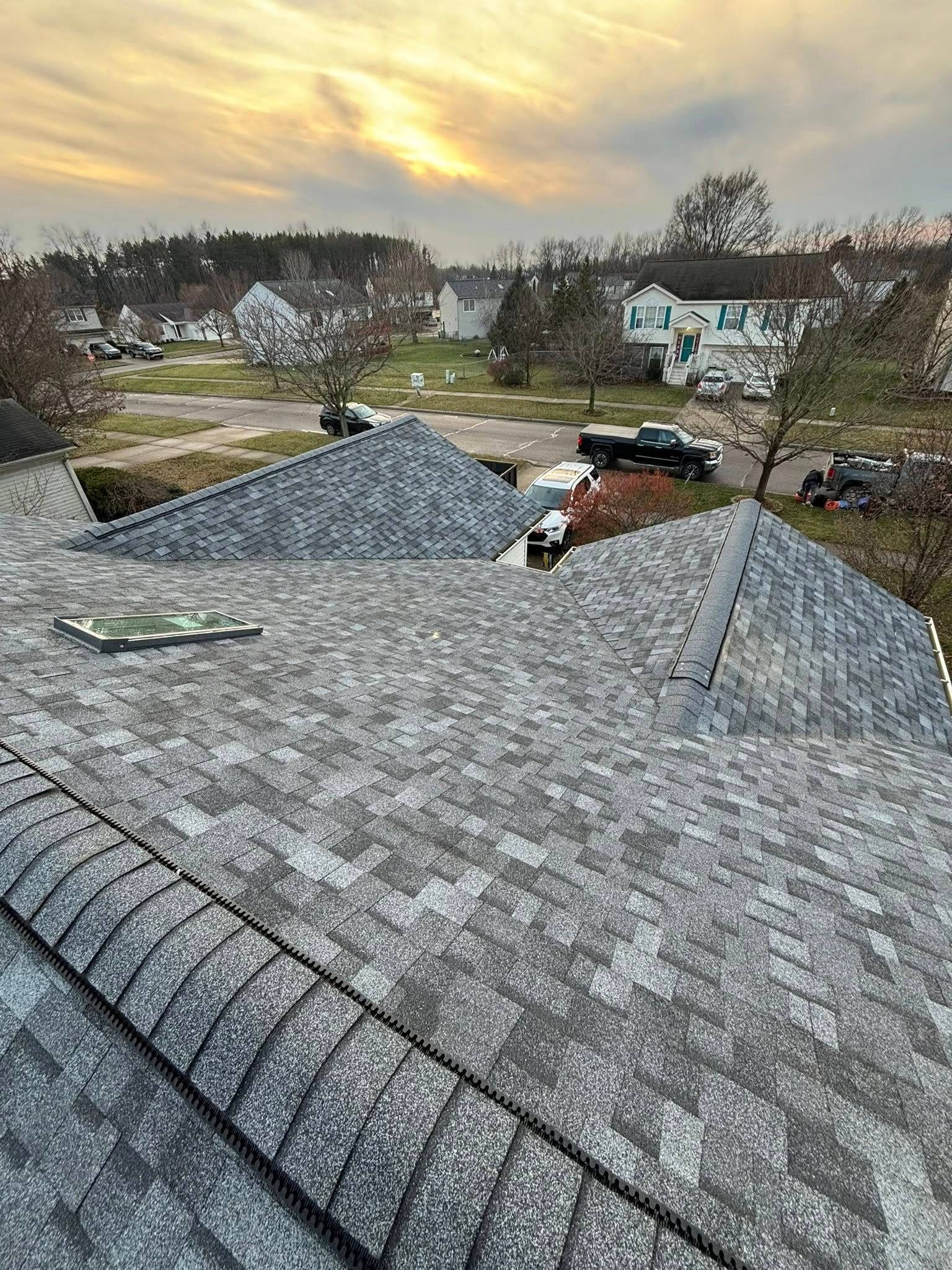 View from a roof, featuring gray shingles. Trees, cars, and buildings are in the background, with a colorful sunset sky.
