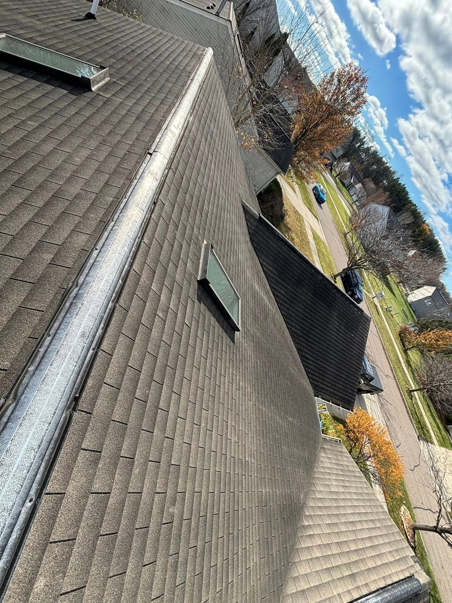 Overhead view of a gray asphalt shingle roof with a ridge vent. Trees and blue sky with clouds in the background.