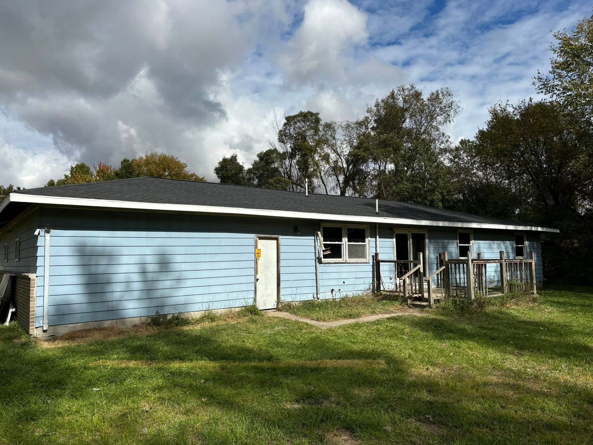 Blue house with weathered siding and overgrown yard under a cloudy sky.