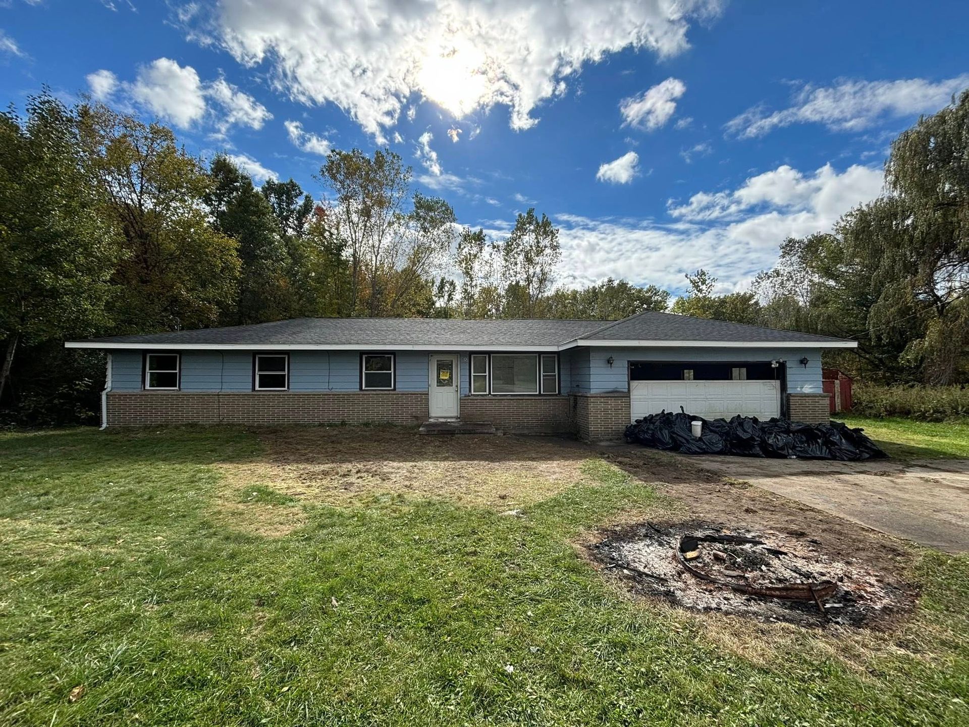 Blue ranch-style house with a gray roof and attached garage, in a yard with grass and trees under a cloudy sky.