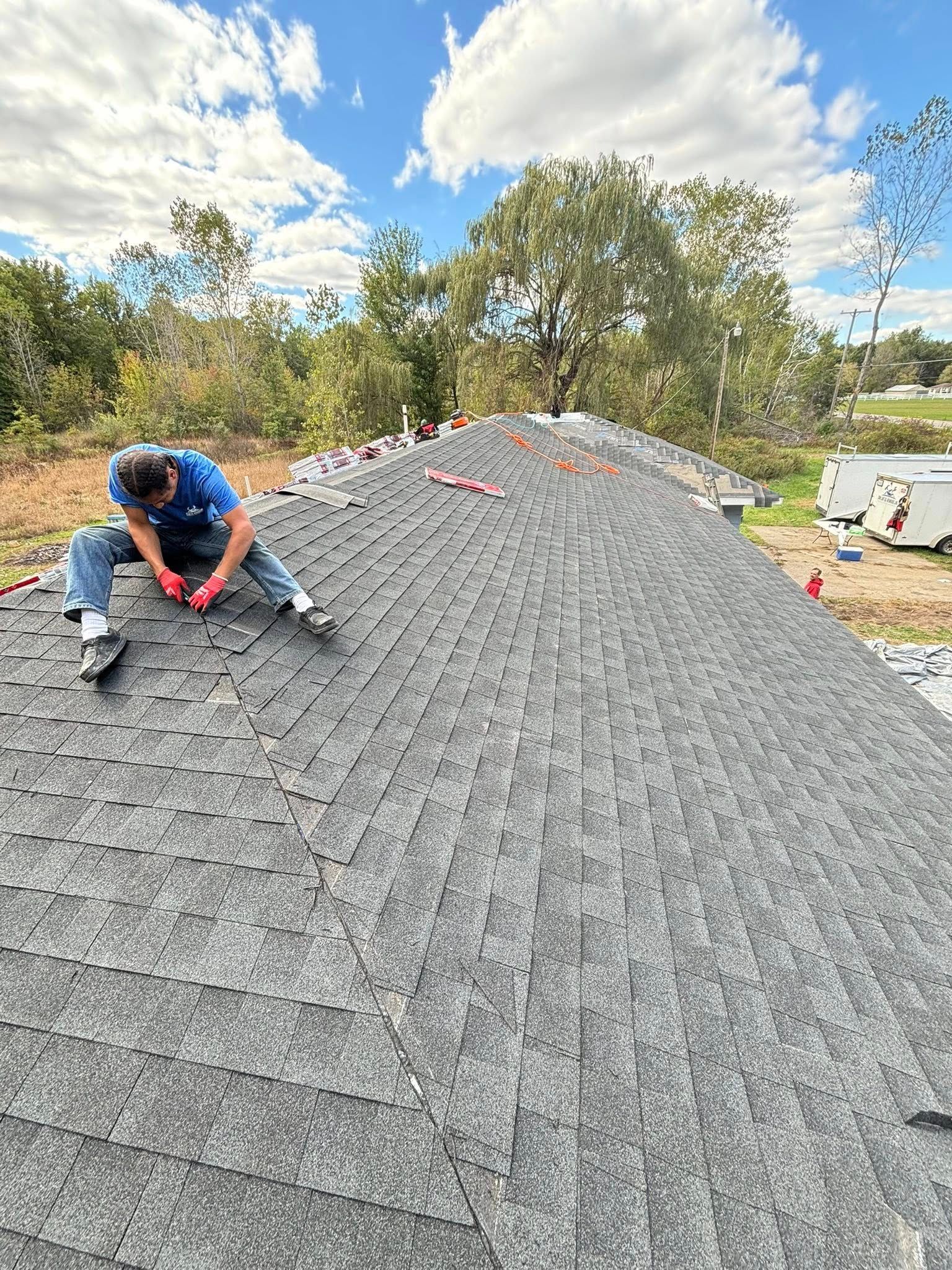 Roofer on a gray shingle roof under a blue sky, installing shingles, wearing work clothes and gloves.