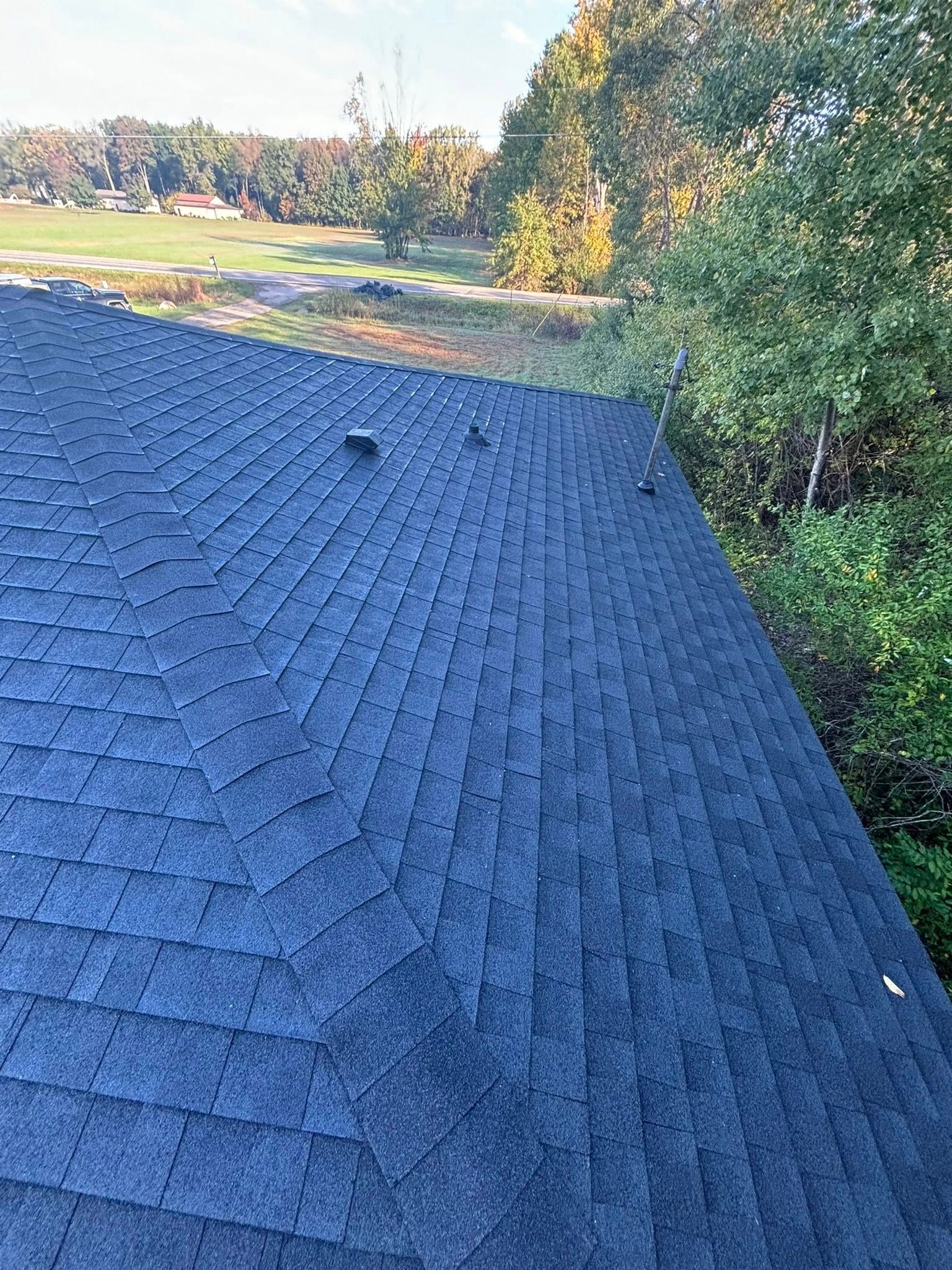 Dark blue asphalt shingle roof, with ventilation stacks, set against a background of trees and a field.