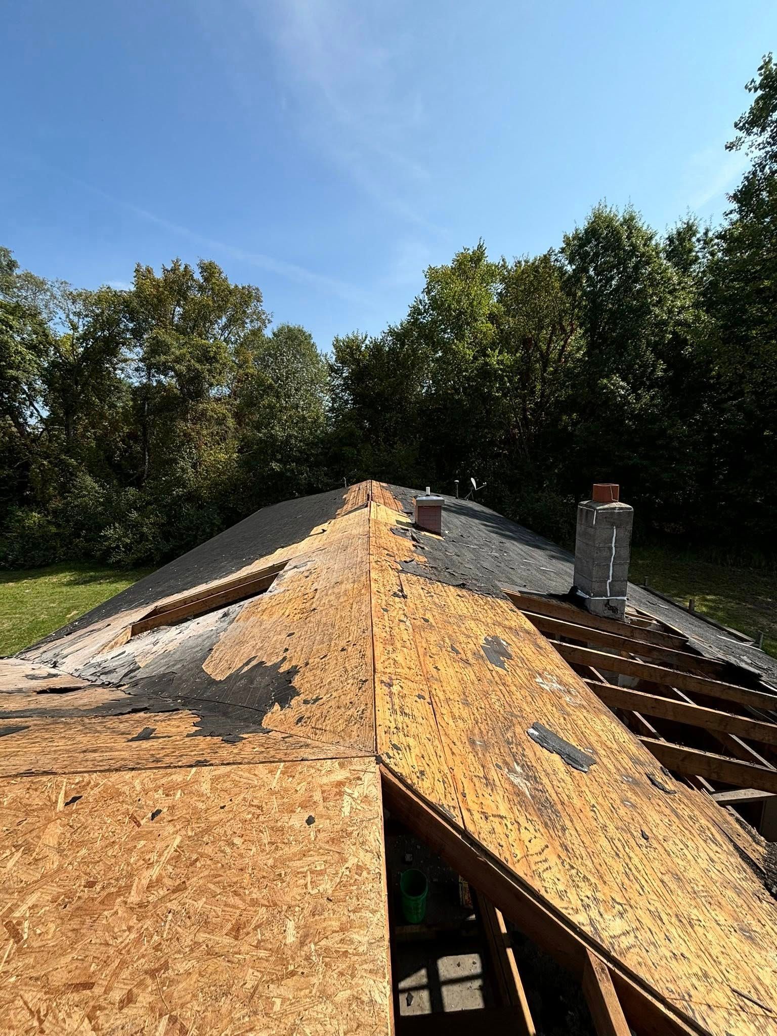 View of a partially stripped house roof, revealing plywood and a chimney against a blue sky.