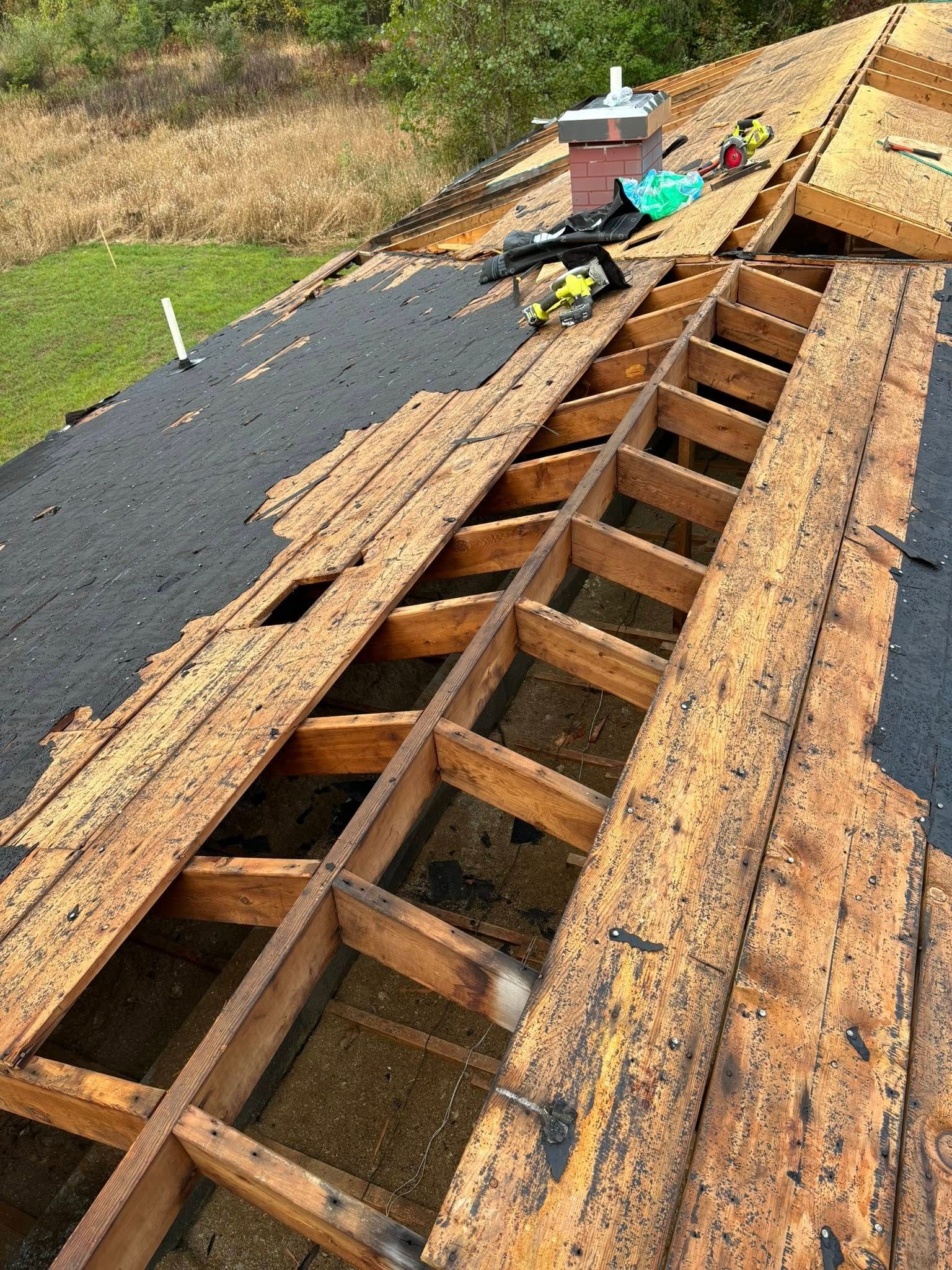 Roof undergoing repair; exposed wooden rafters and decking. Old shingles removed, tools and materials present.