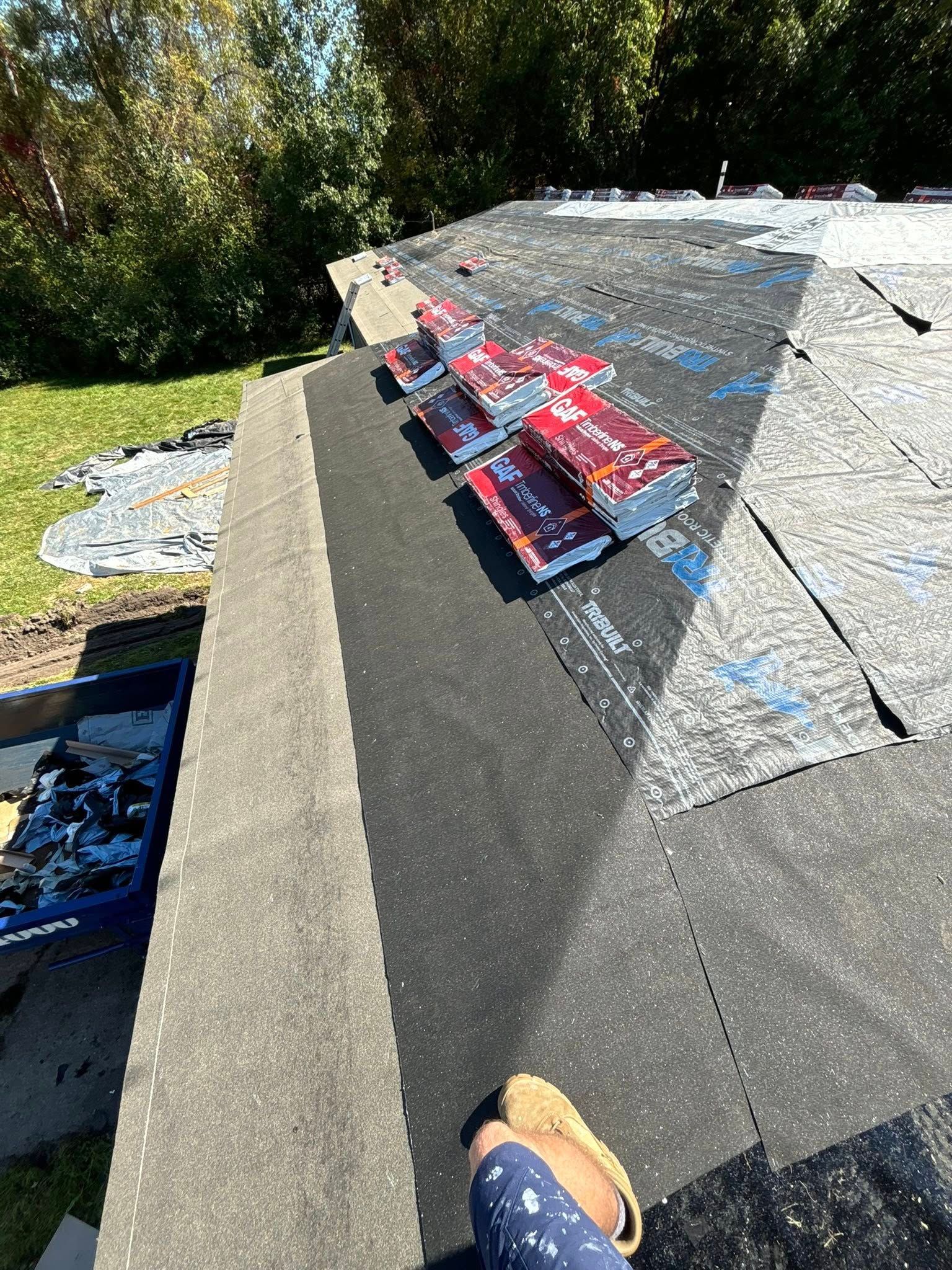 Rooftop with shingles and tools; person's foot in foreground. Sunny day, green trees in the background.