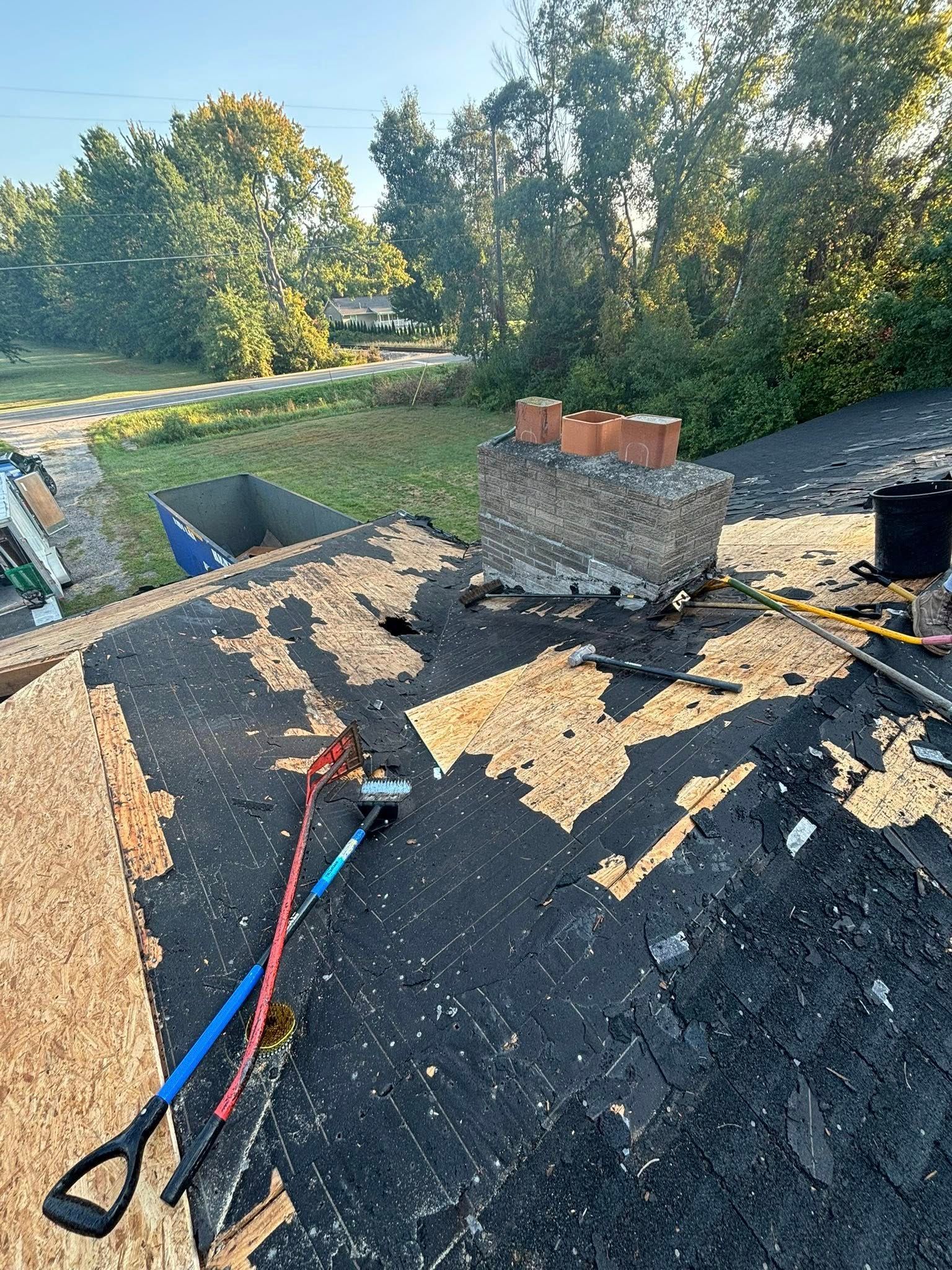 A damaged roof with a brick chimney, tools, and trees in the background.