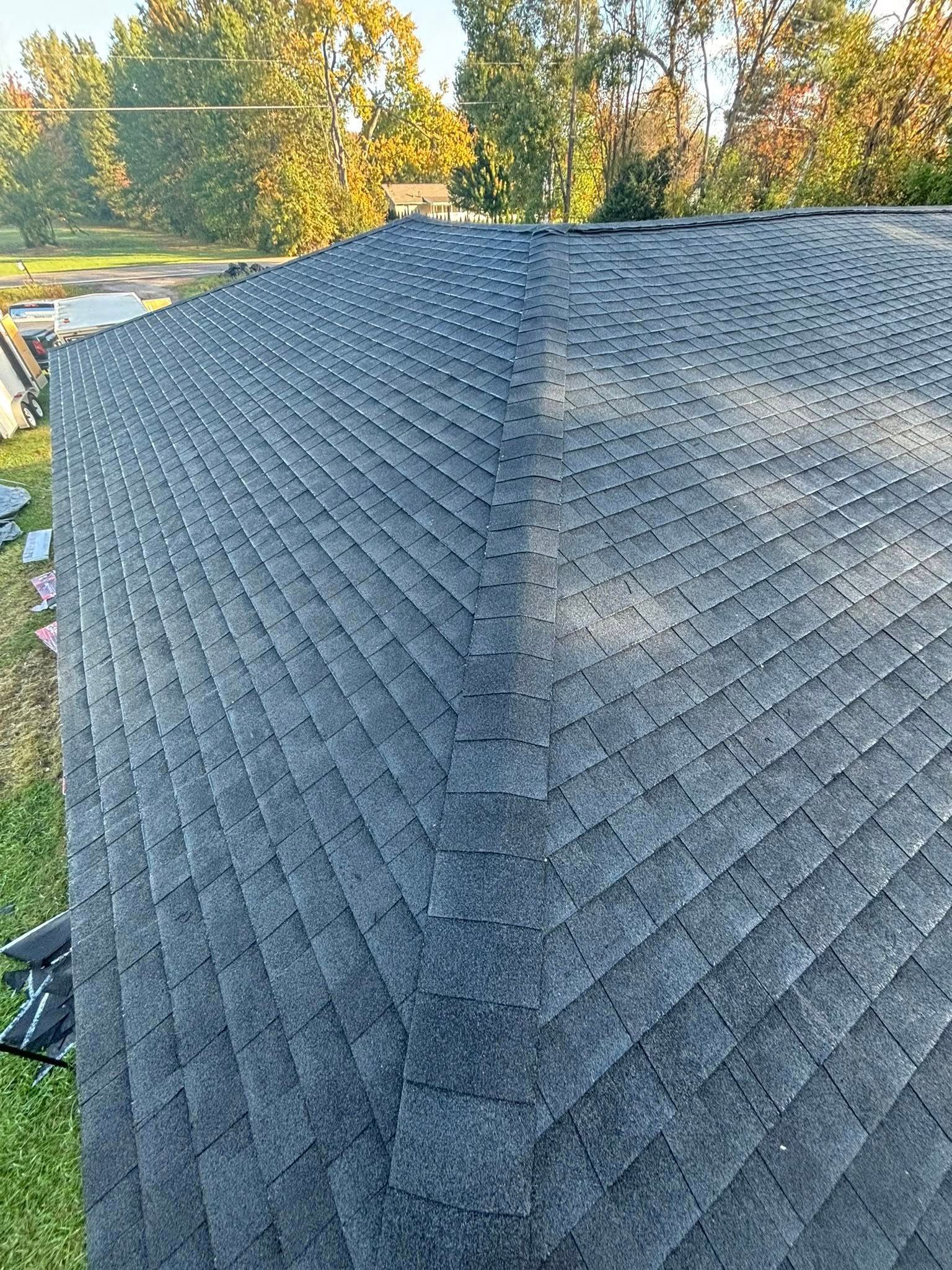 Dark gray asphalt shingle roof. The roof has a sharp corner, with trees and sky in the background.