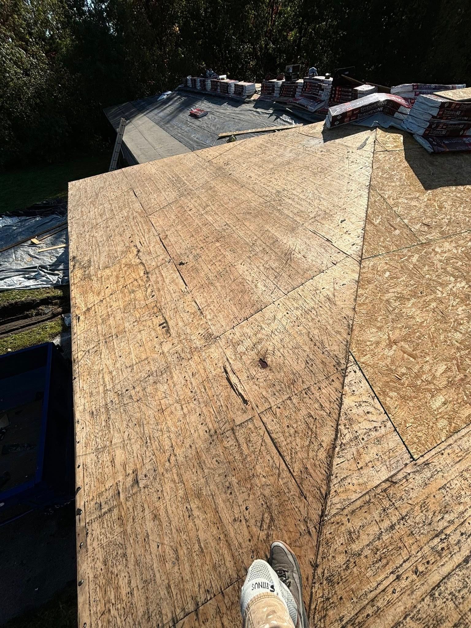 New plywood roof surface with stacked shingles in the background; a person's foot is visible in the foreground.