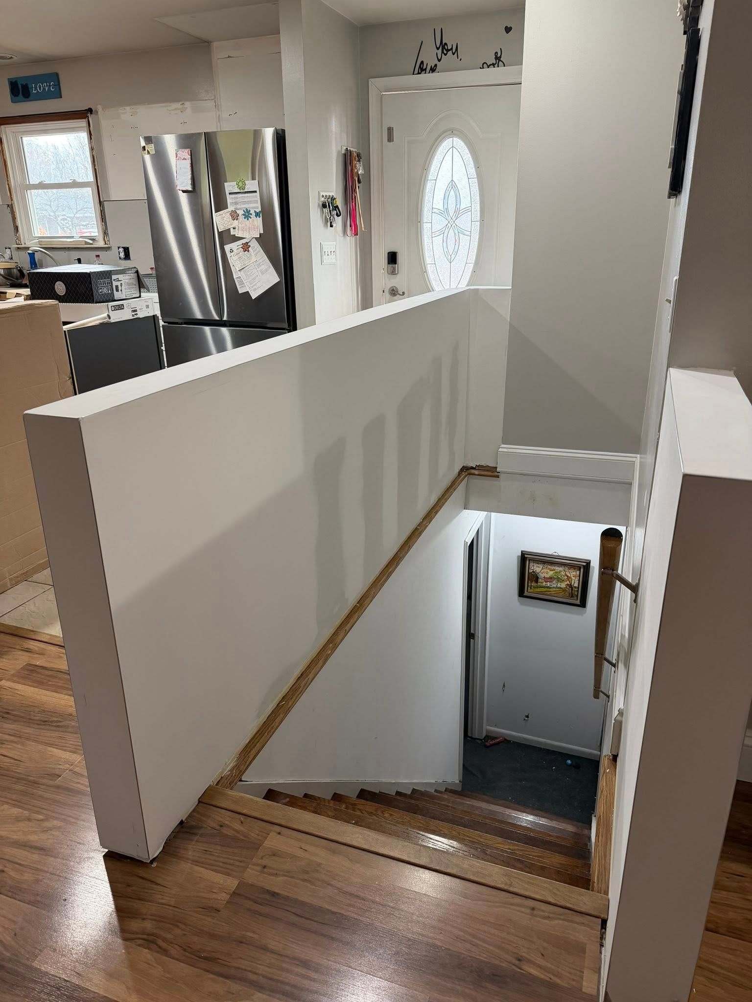 View of stairs leading down, with white wall and wooden floor. Kitchen visible in the background.