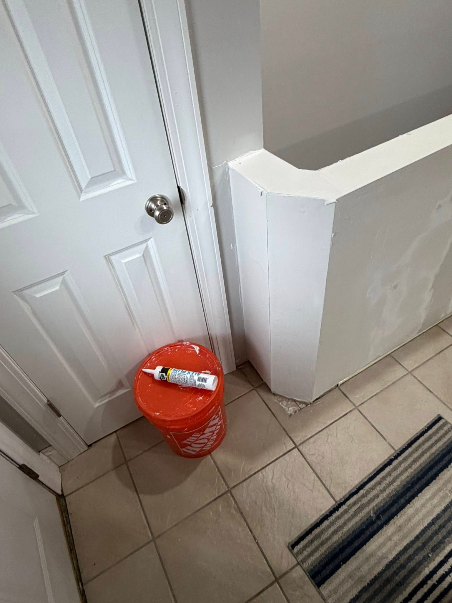 Orange bucket with caulk tube next to a white door and wall. Tile floor, rug.