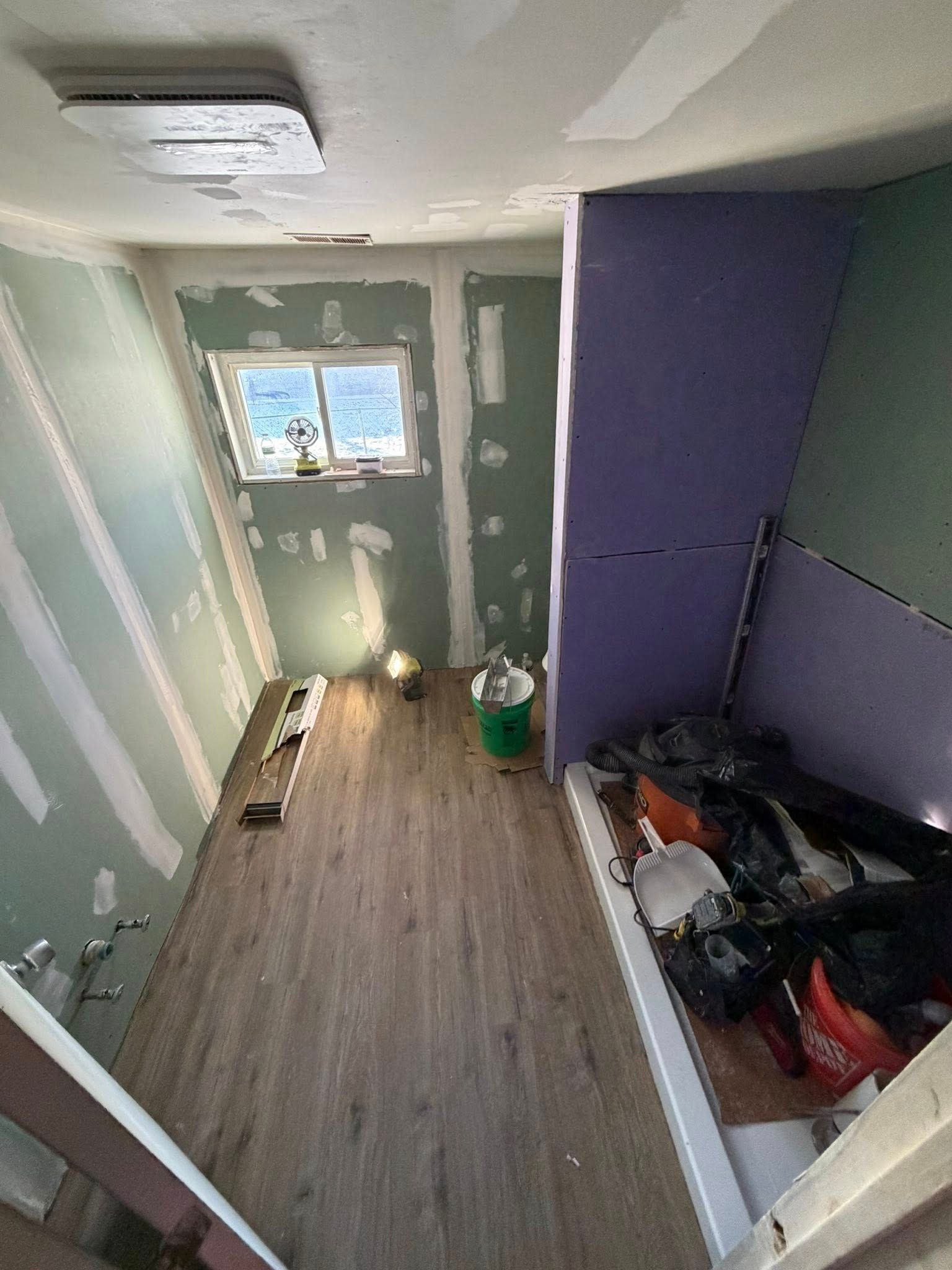 Bathroom under renovation; walls with green and purple drywall; wooden floor. Construction supplies visible.
