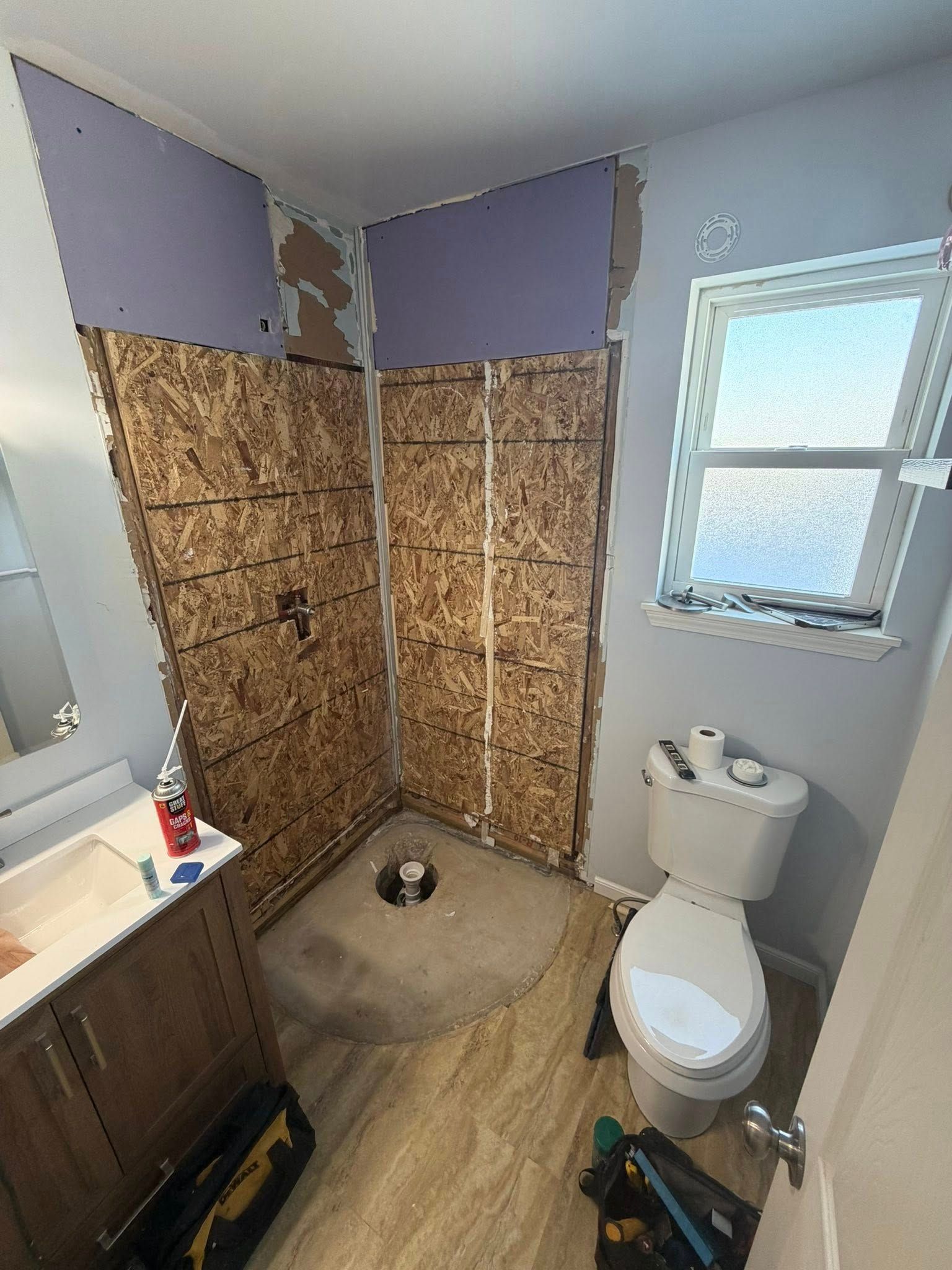 Bathroom under renovation: shower with wooden paneling, toilet, and vanity. Purple drywall and window visible.