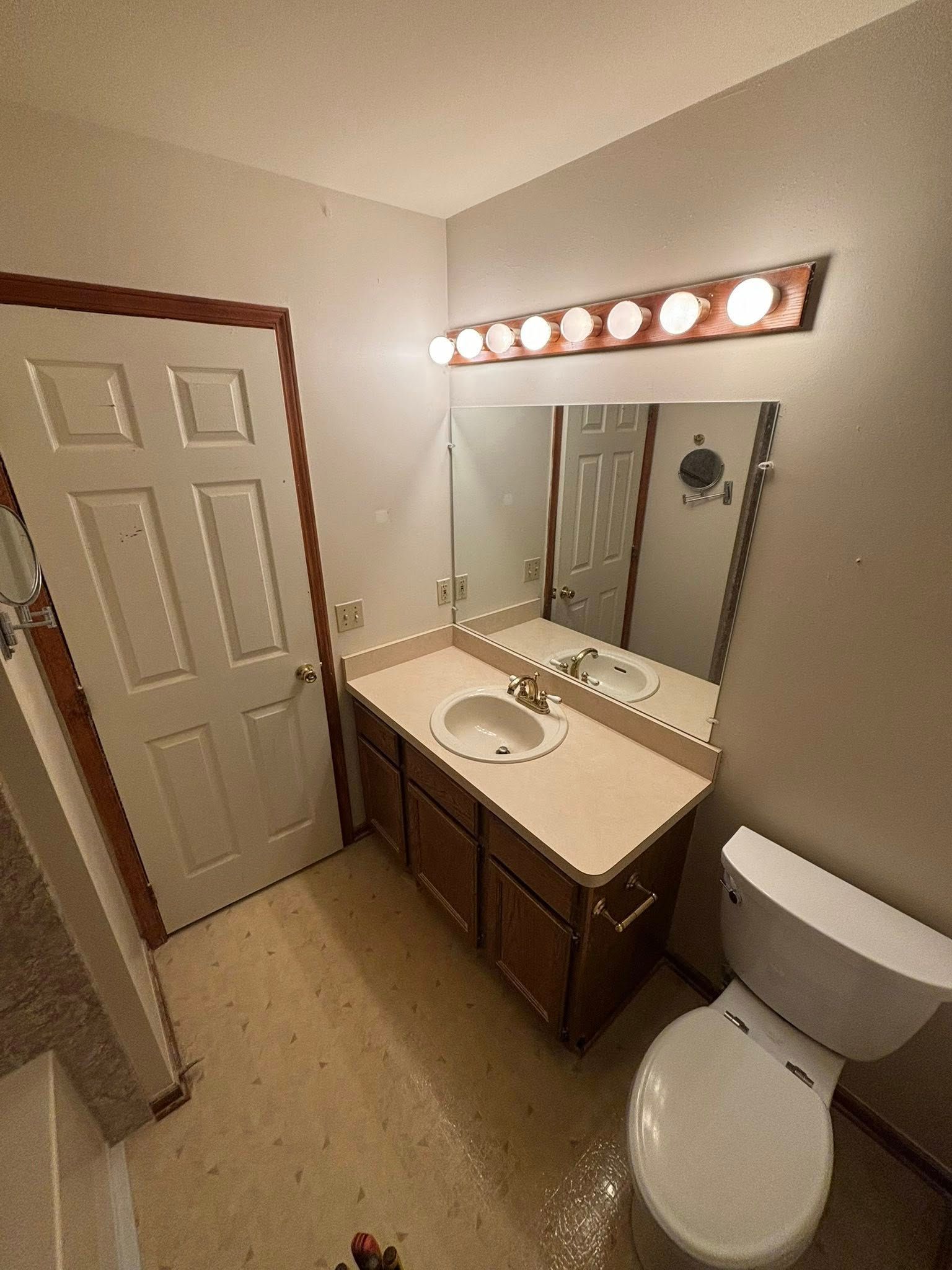 Bathroom with a vanity, toilet, and door. Neutral walls, brown cabinets and door frame.