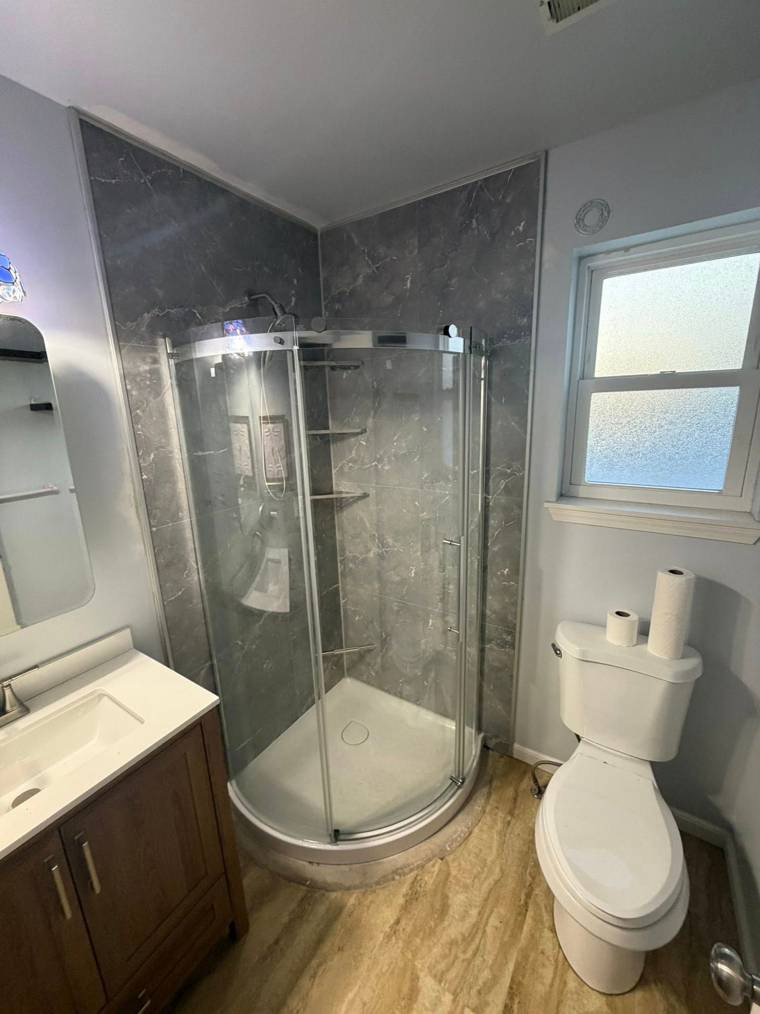 Bathroom with a curved glass shower, toilet, and vanity. Gray tile walls and tan flooring.
