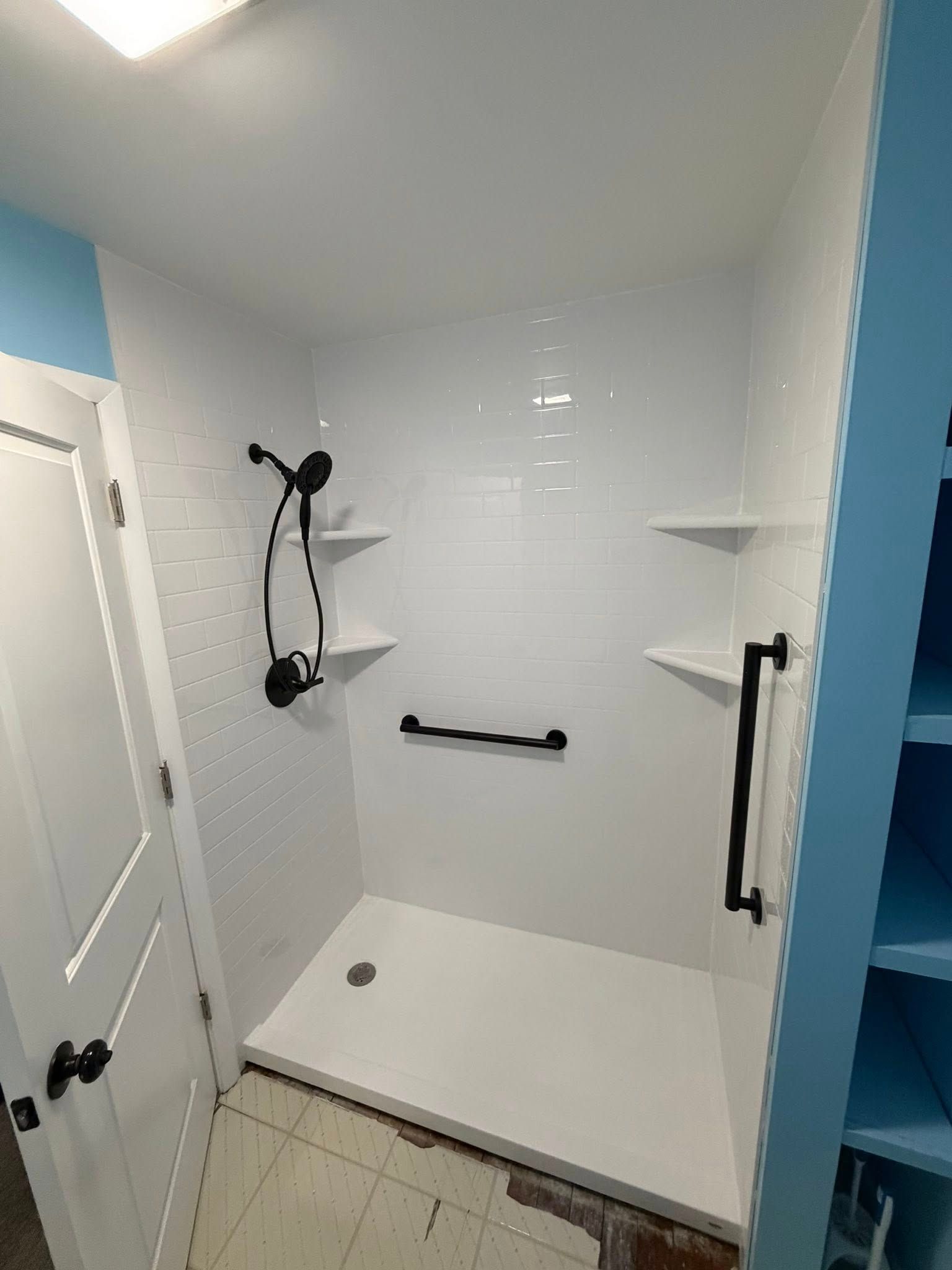White tiled shower with black fixtures and grab bars, two shelves, and door on the left.