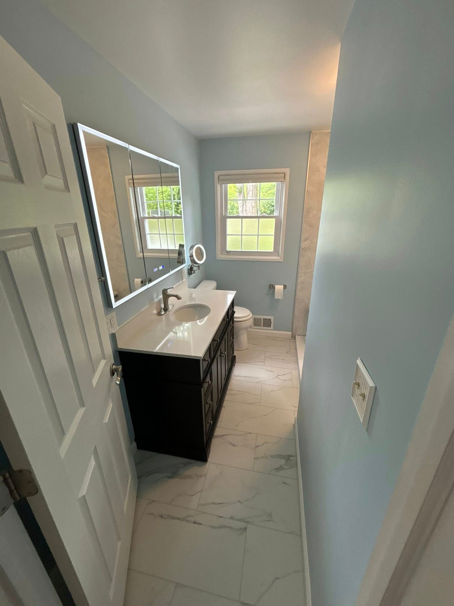Bathroom with blue walls, dark vanity, marble floor, lighted mirror, and window.