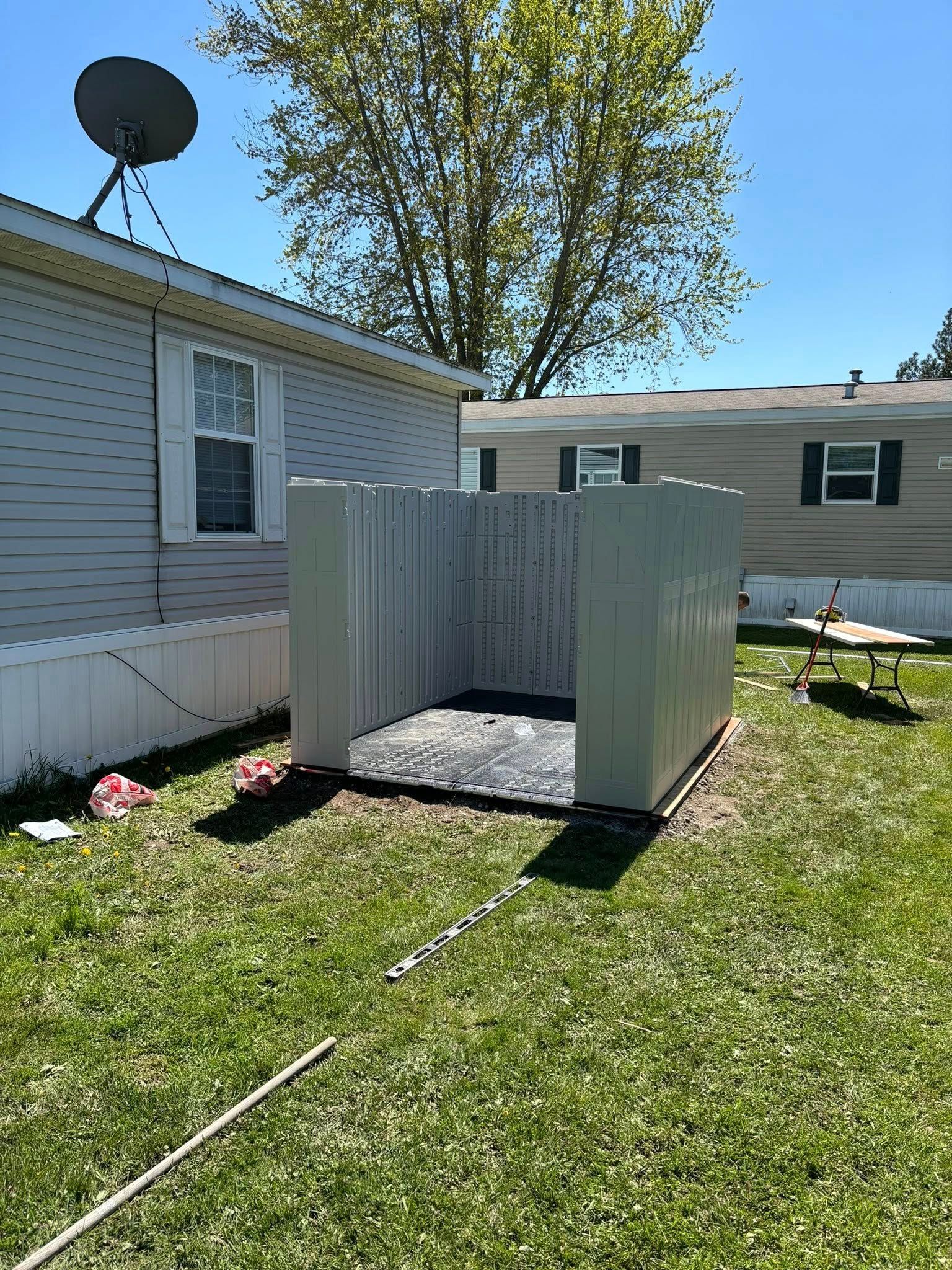 A gray shed-like structure sits in a grassy yard, next to a mobile home, under a sunny sky.