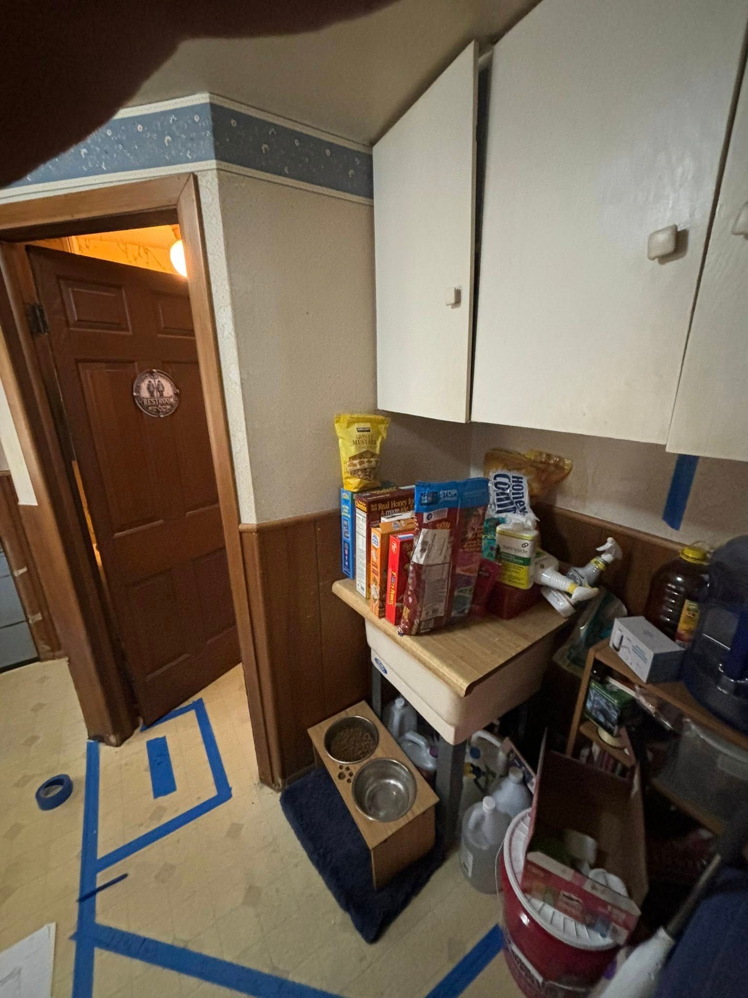 A cluttered pantry area with pet food and supplies on a small table, cabinets above, and a door on the left.