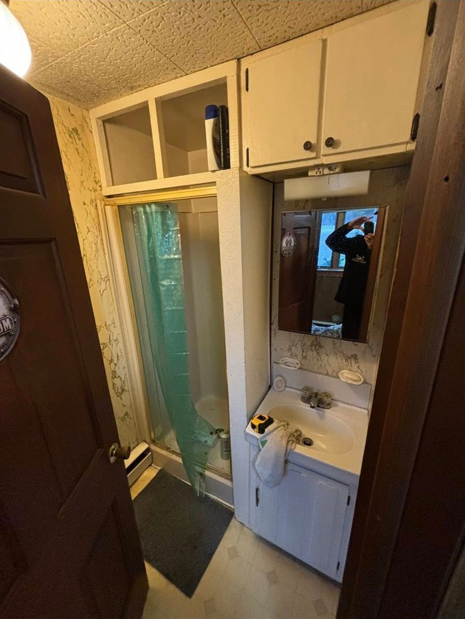 Small bathroom with a shower, sink, and overhead cabinets; a person is reflected in the mirror.