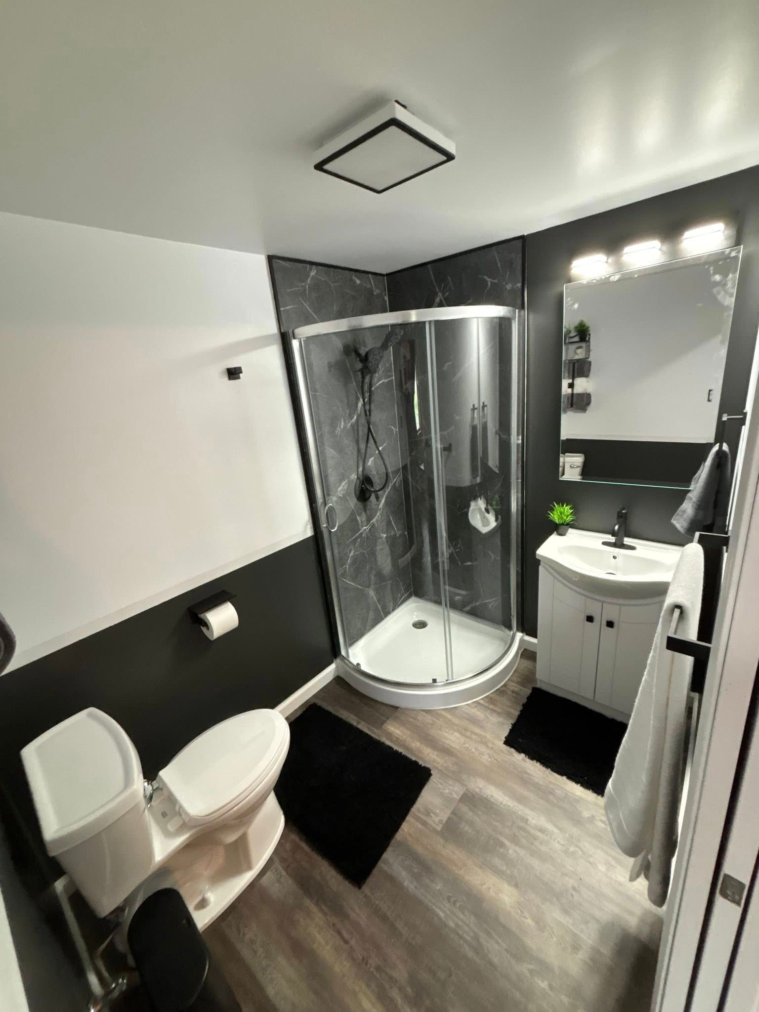 Modern bathroom with black and white accents: shower, toilet, vanity, black rugs, wood-look floor.