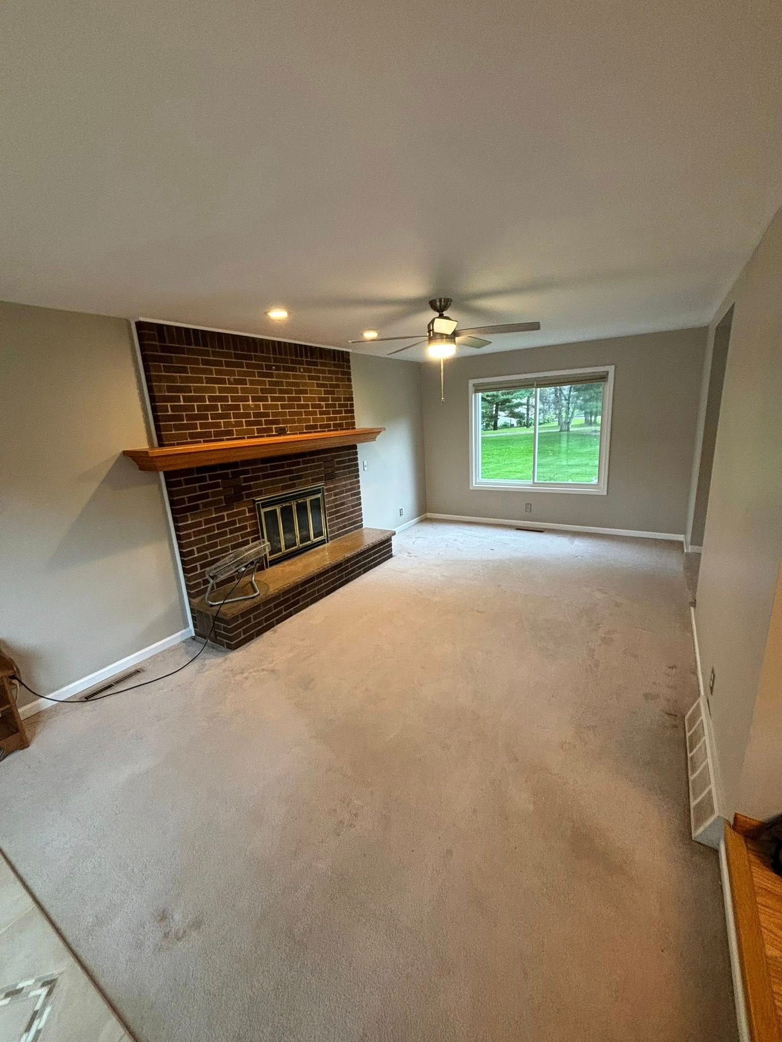 Living room with a brick fireplace and a window, beige carpet, and gray walls.