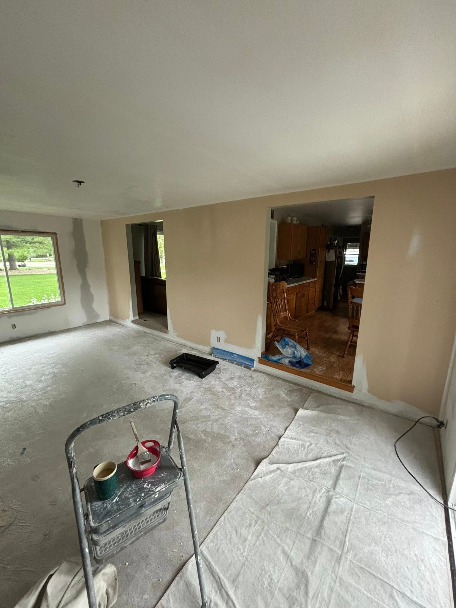 Room under renovation; beige walls, bare floors, open doorways. Paint supplies on ladder.