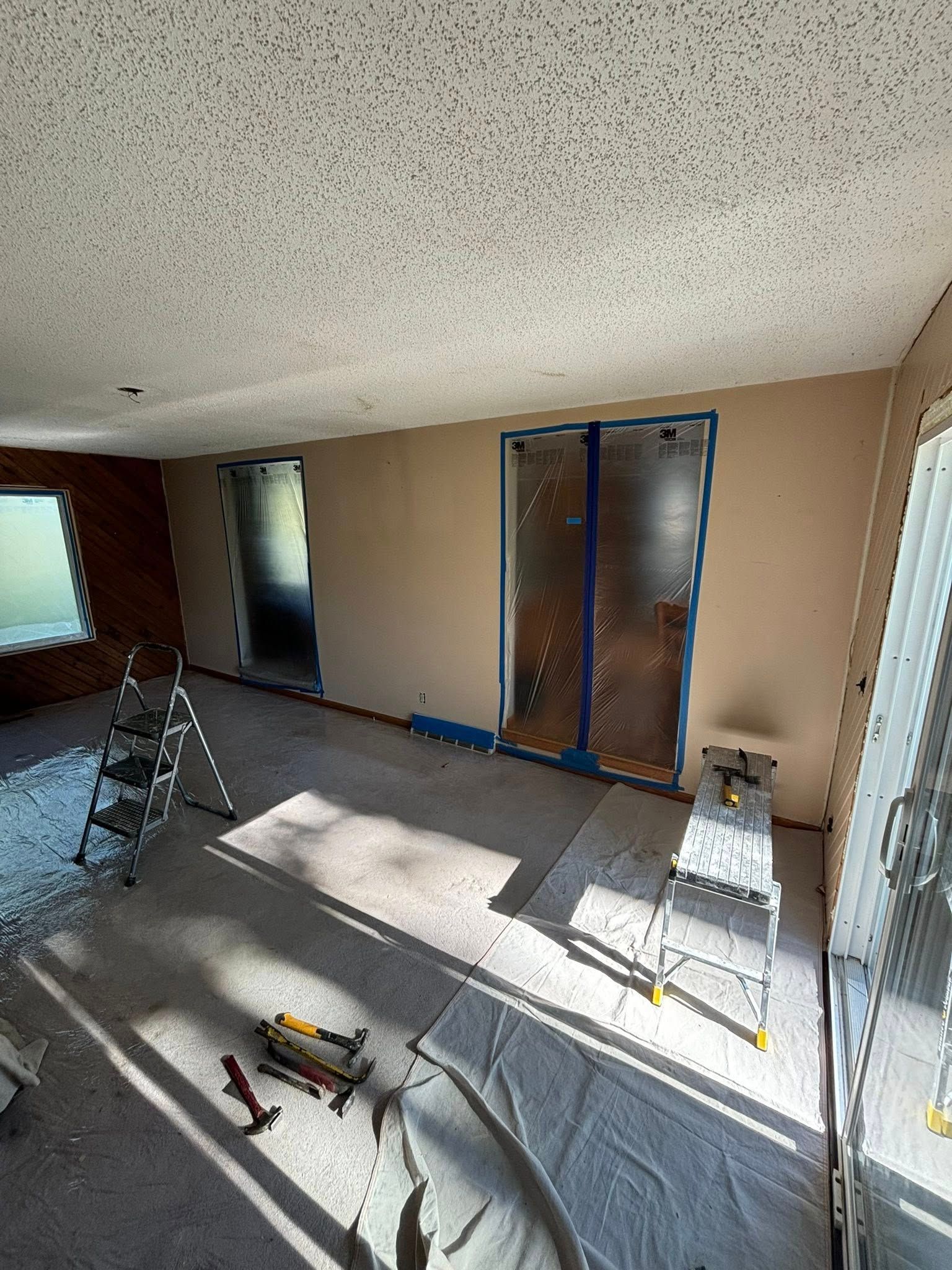 Room with tan walls, taped-off openings, ladders, and protective sheeting, indicating a painting project in progress.