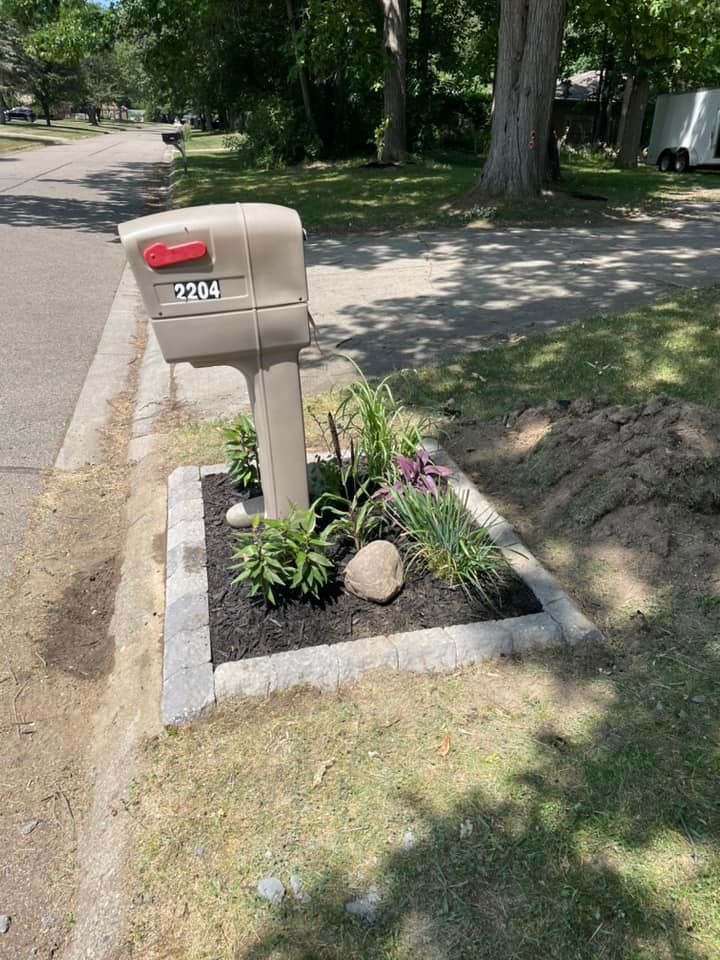 Beige mailbox with red flag sits above a landscaped bed with dark mulch, plants, and a large stone.