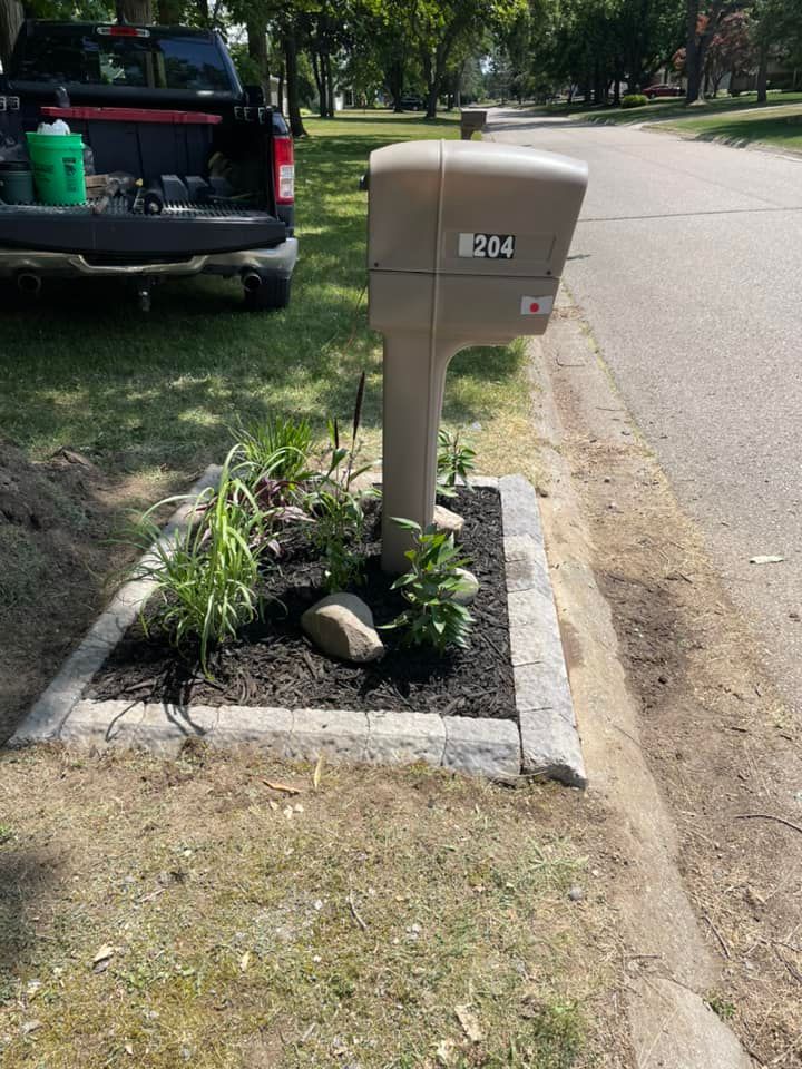 Beige mailbox surrounded by a small garden bed with rocks, mulch, and plants next to a road.
