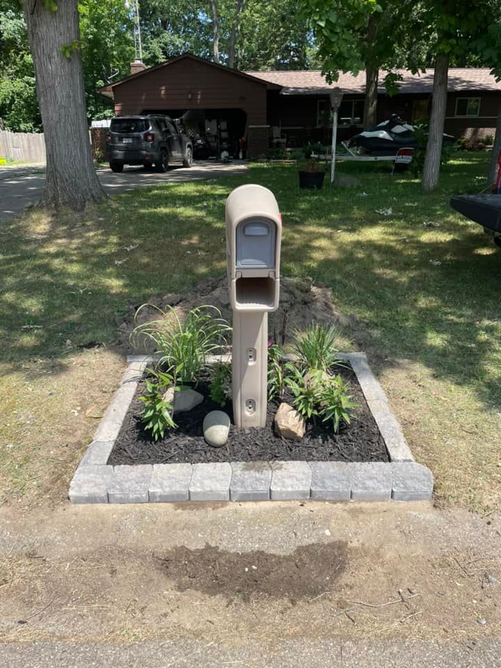 Beige mailbox with small garden bed and dark mulch, set in a gray brick border, in front of a house.