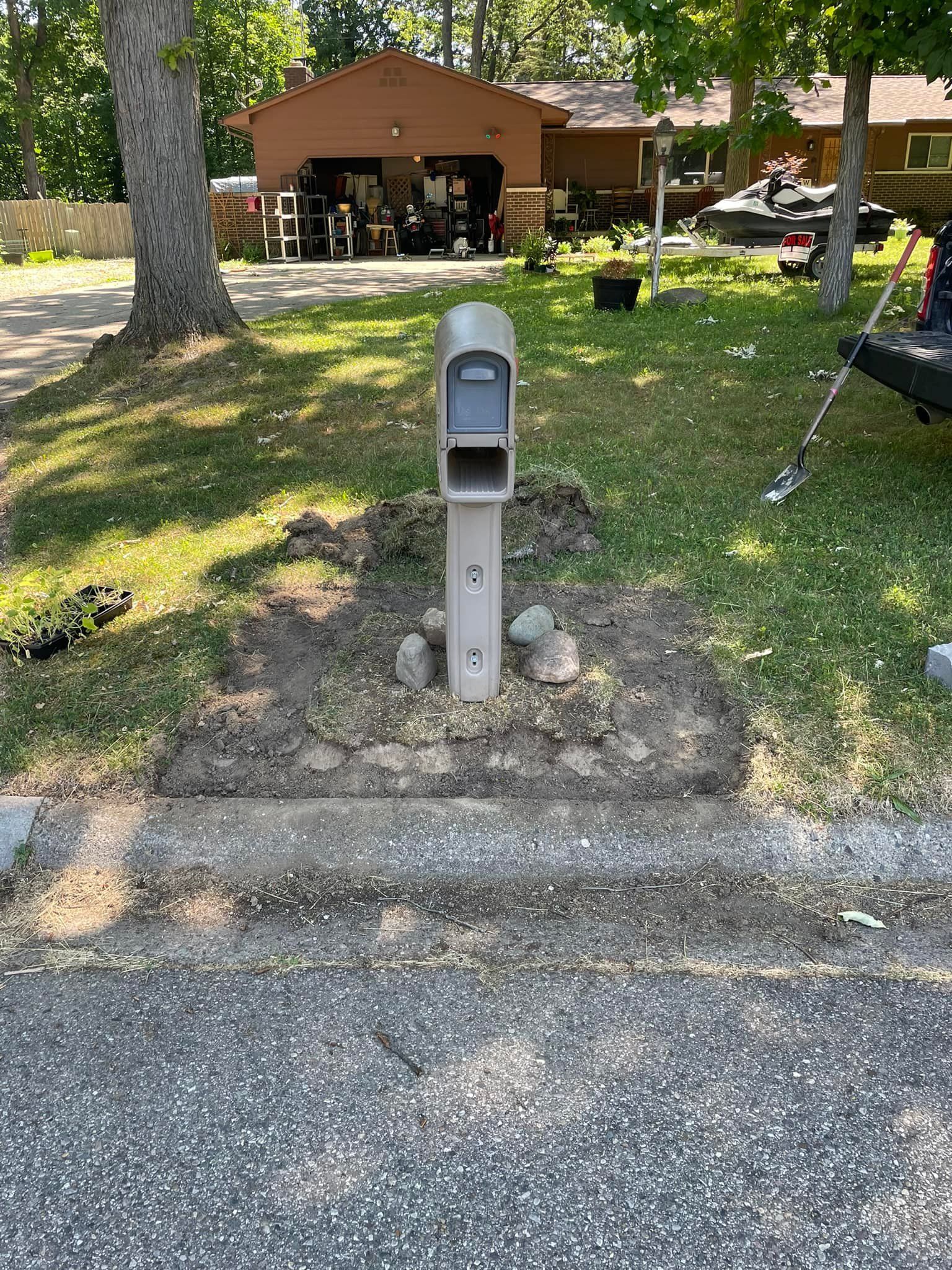 A beige mailbox on a post sits in a cleared area next to a paved road.