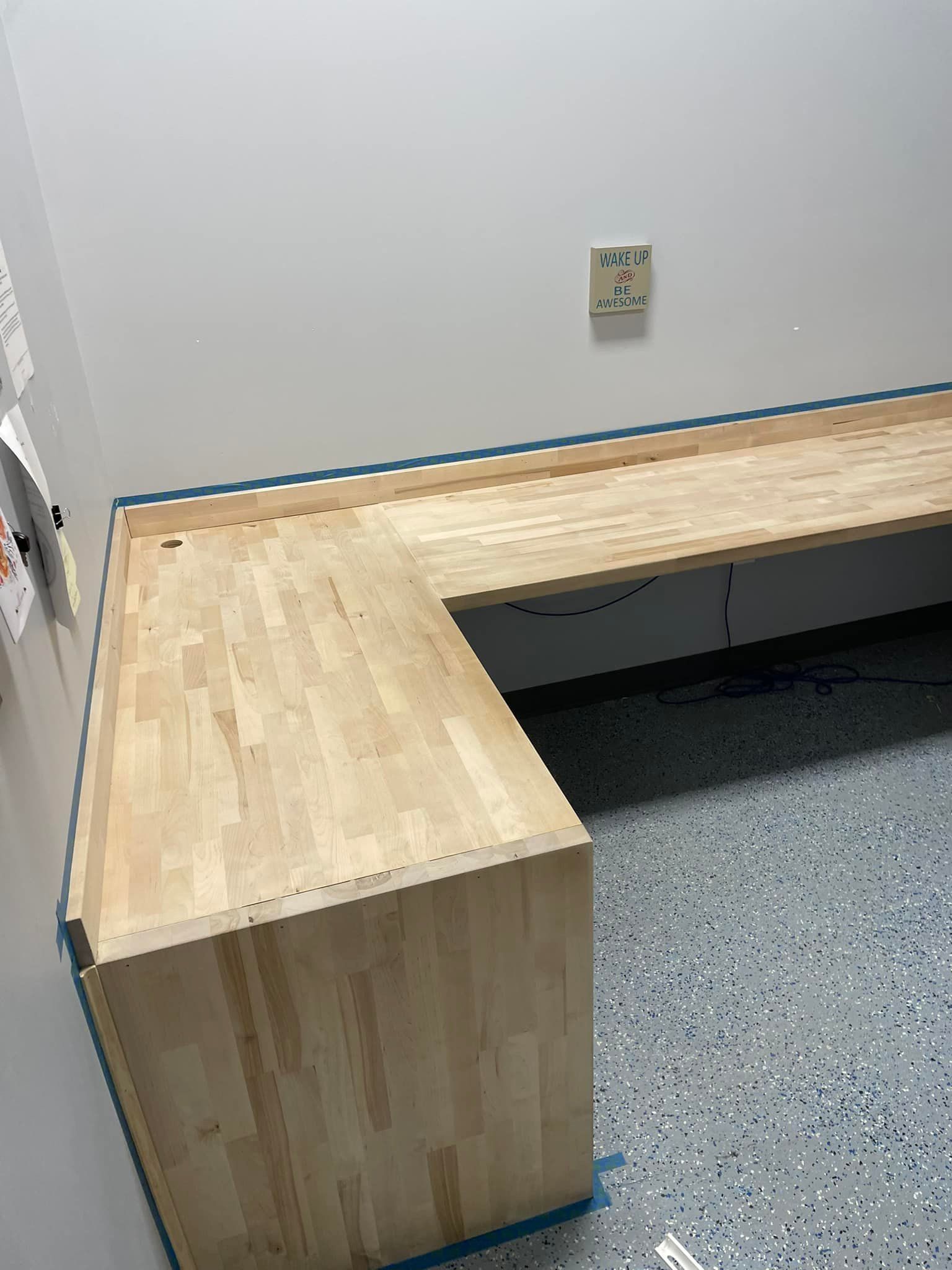 A light-colored butcher block desk in a corner against a white wall and blue speckled floor.