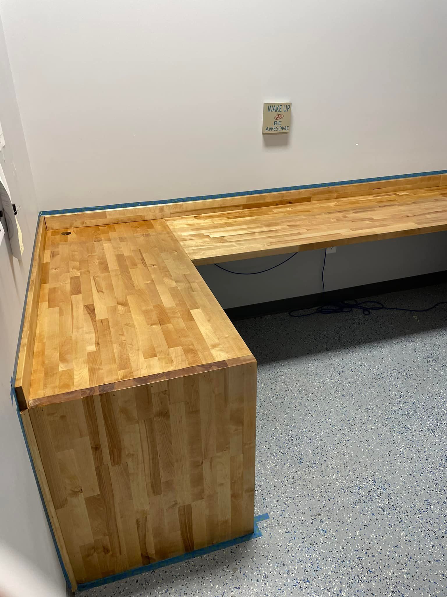 L-shaped wooden workbench against white wall, with a speckled floor and an electrical outlet.