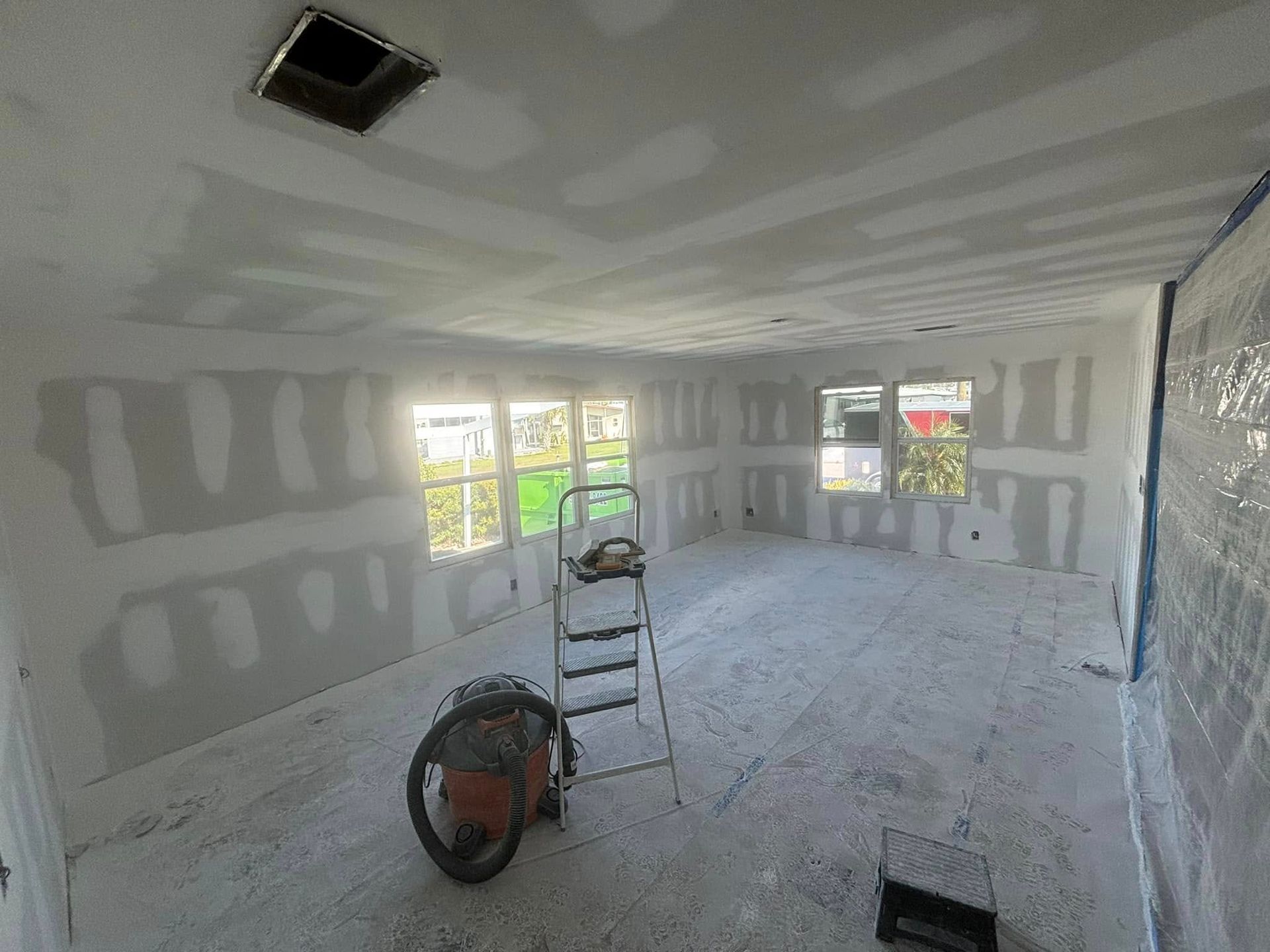 Interior room under construction, with drywall installed on walls and ceiling. Ladder, fan, and tools present.