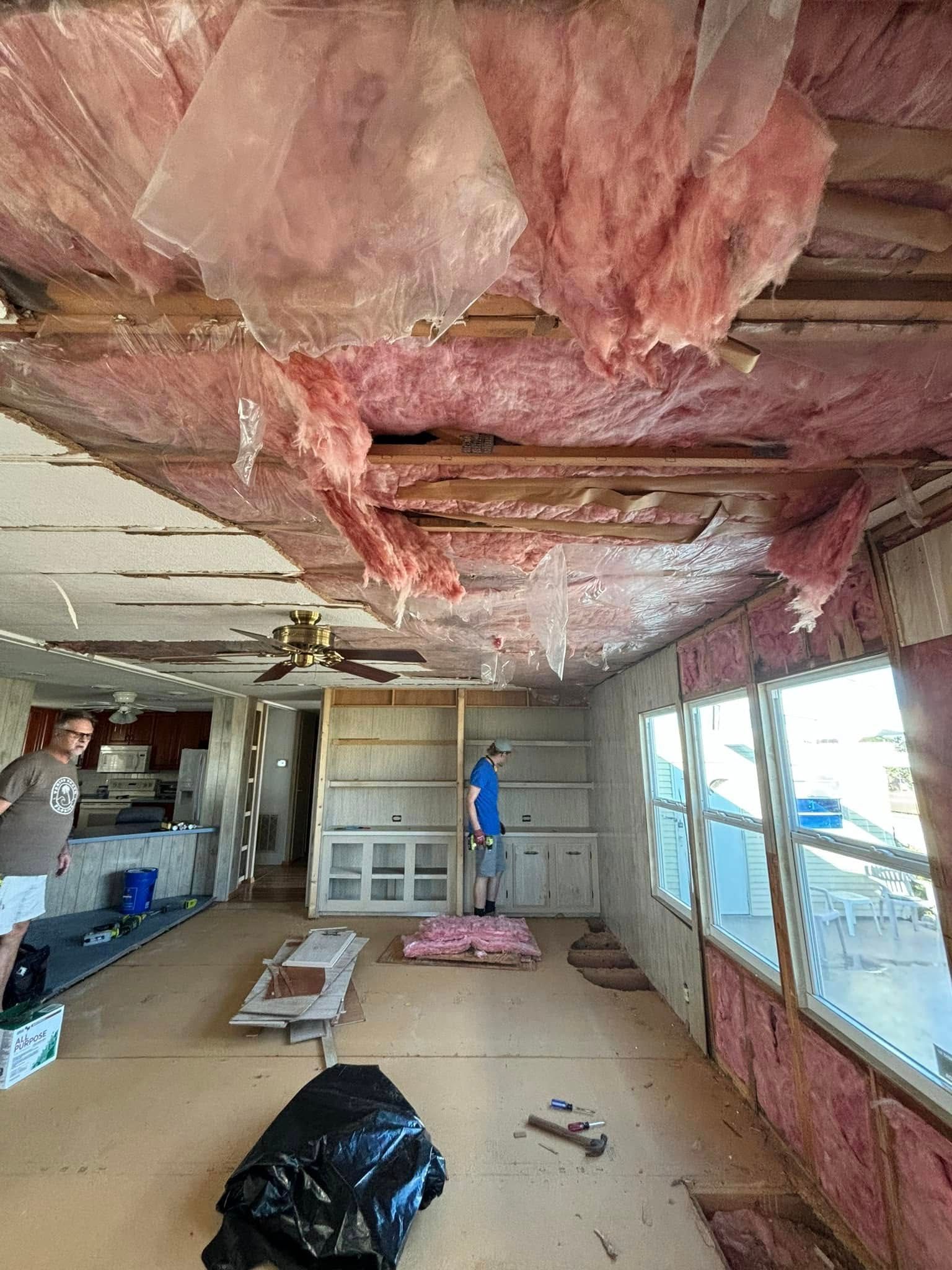 Interior room undergoing renovation; exposed ceiling insulation. Two people inside. Pink insulation, wood, and white walls.