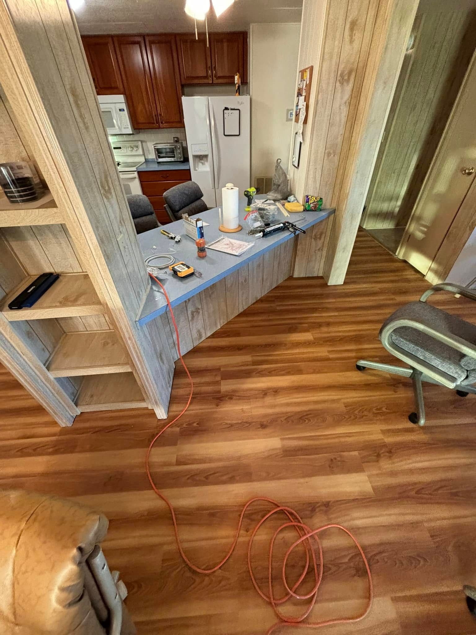 Room with a desk, shelves, and kitchen visible. A red extension cord runs across a wood-look floor.