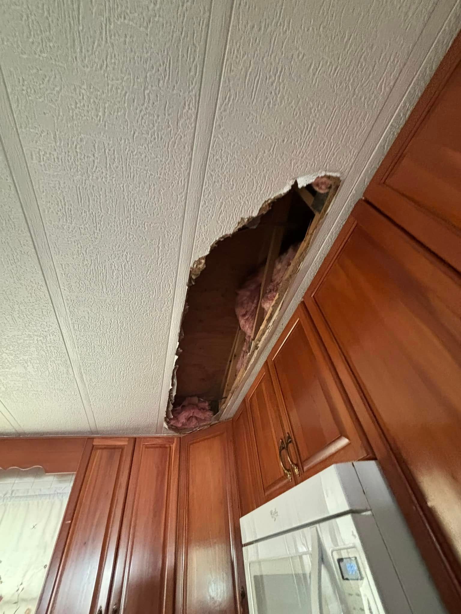 Hole in textured ceiling above wooden cabinets, exposing insulation and wiring, likely kitchen.
