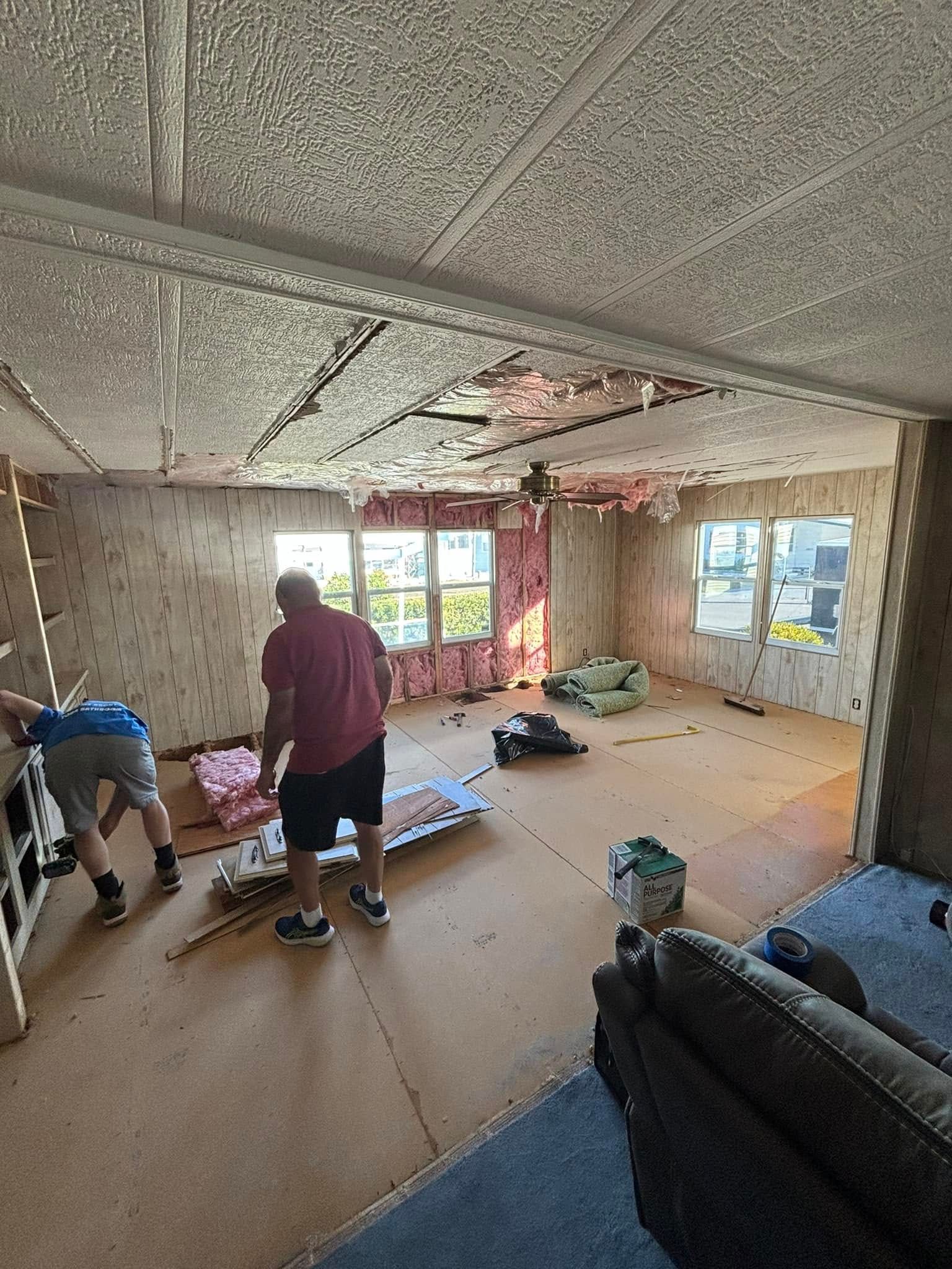 Men renovating a room, ceiling and wall damage. One holds insulation, other works on a wall. Brown floor, windows.