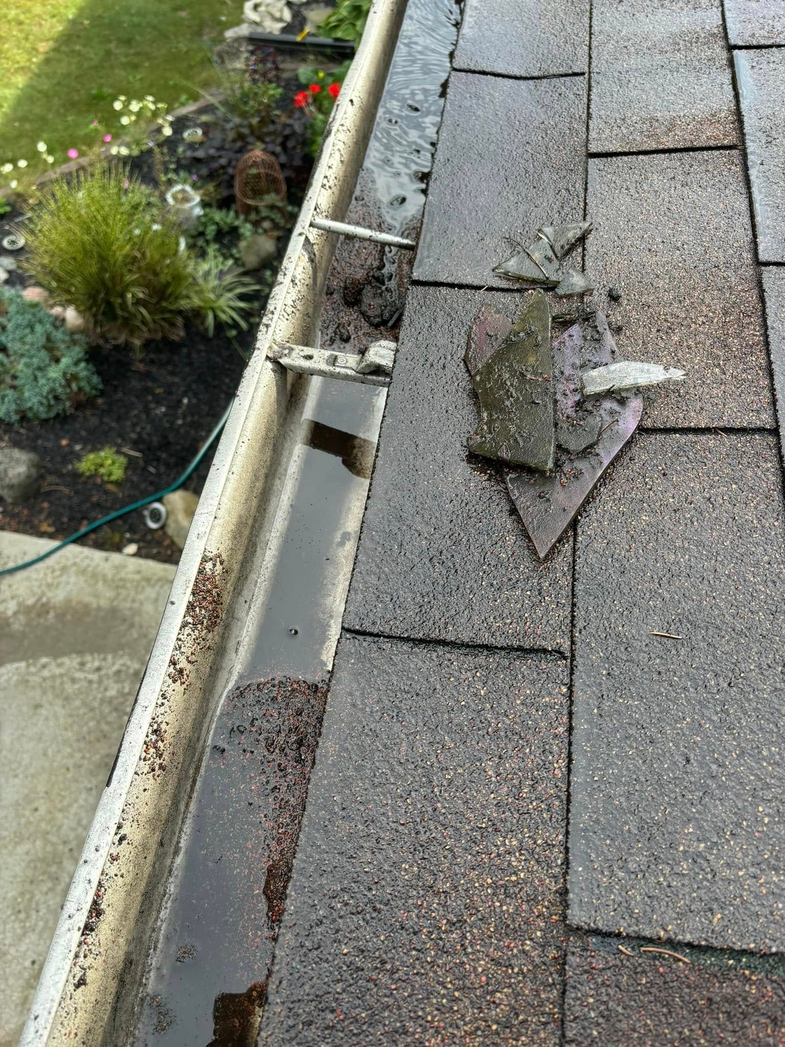 Gutter and roof shingles with water. Shingles are broken near a bracket. Garden in background.