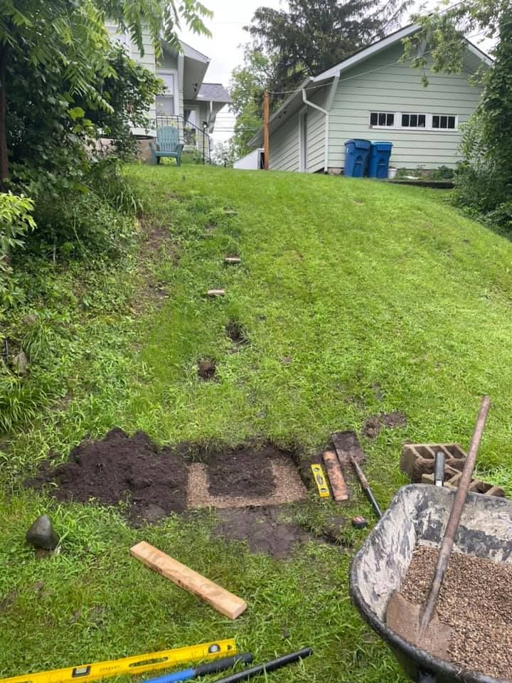 A grassy slope with a path under construction, including steps and tools like shovels, levels, and a wheelbarrow.