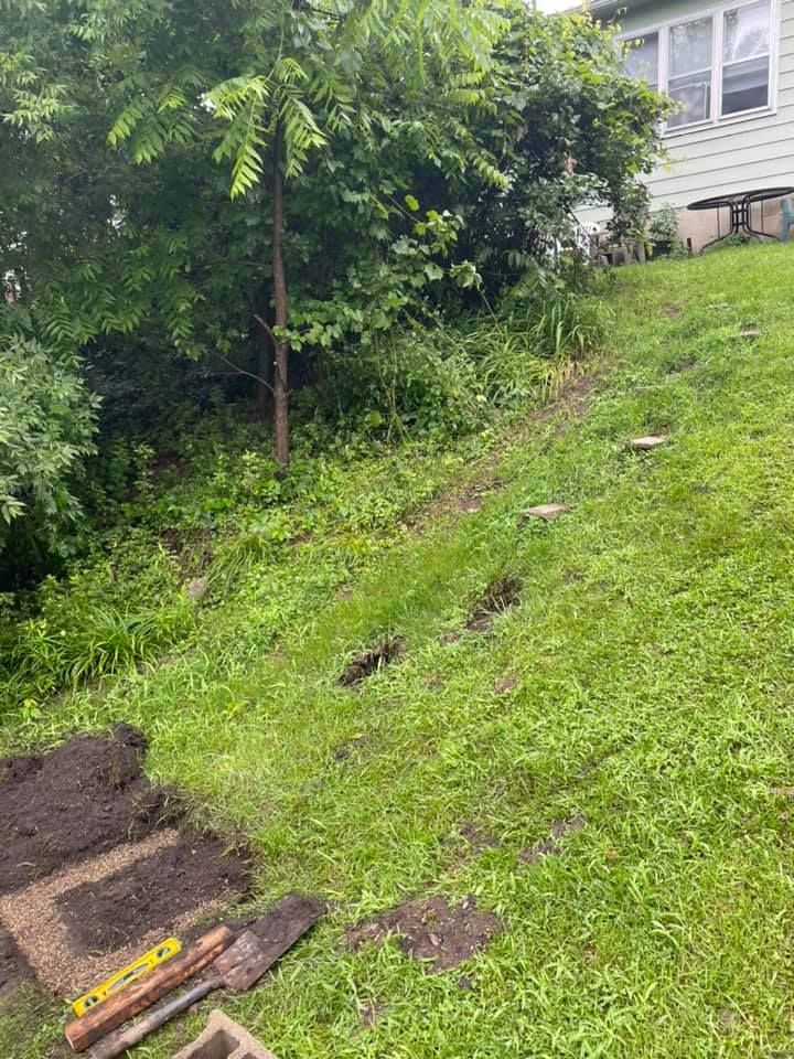 A grassy hillside with steps being built, tools and materials are visible in the foreground near a house.