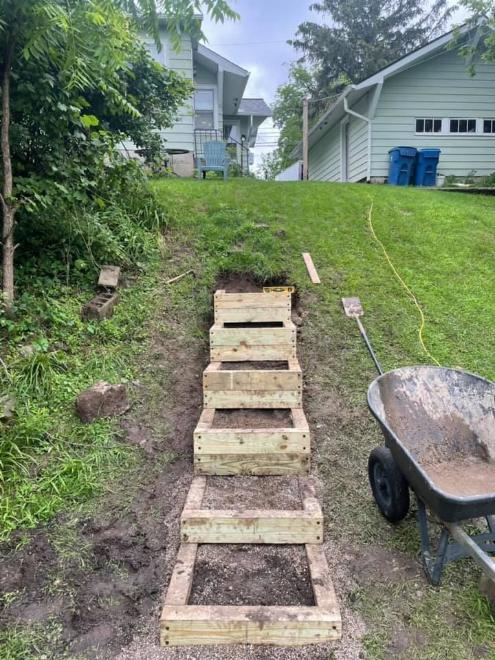 Wooden staircase built into a grassy hillside, leading up to houses; a wheelbarrow and tools are nearby.