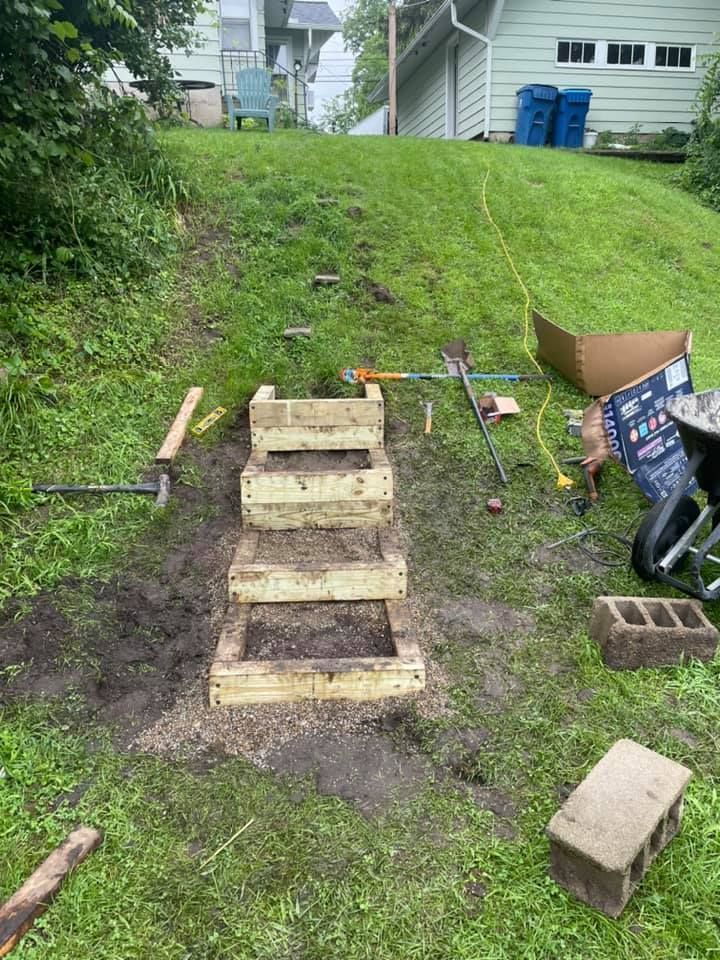 Wooden steps being built on a grassy slope. Tools and materials are scattered around the construction area.
