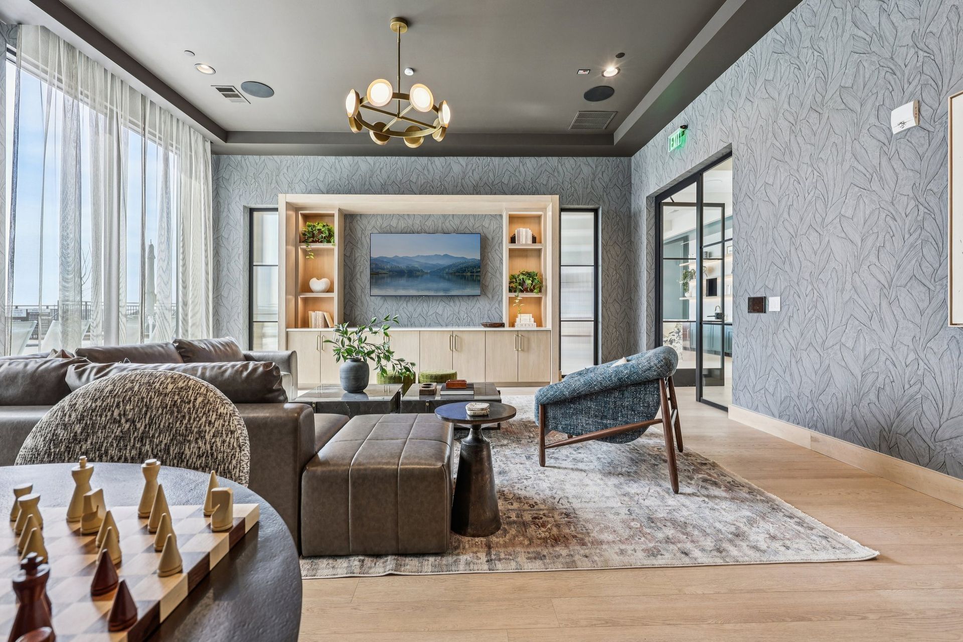 Modern living room with a chess set in the foreground, a gray sectional, patterned wallpaper, and a wooden media unit.