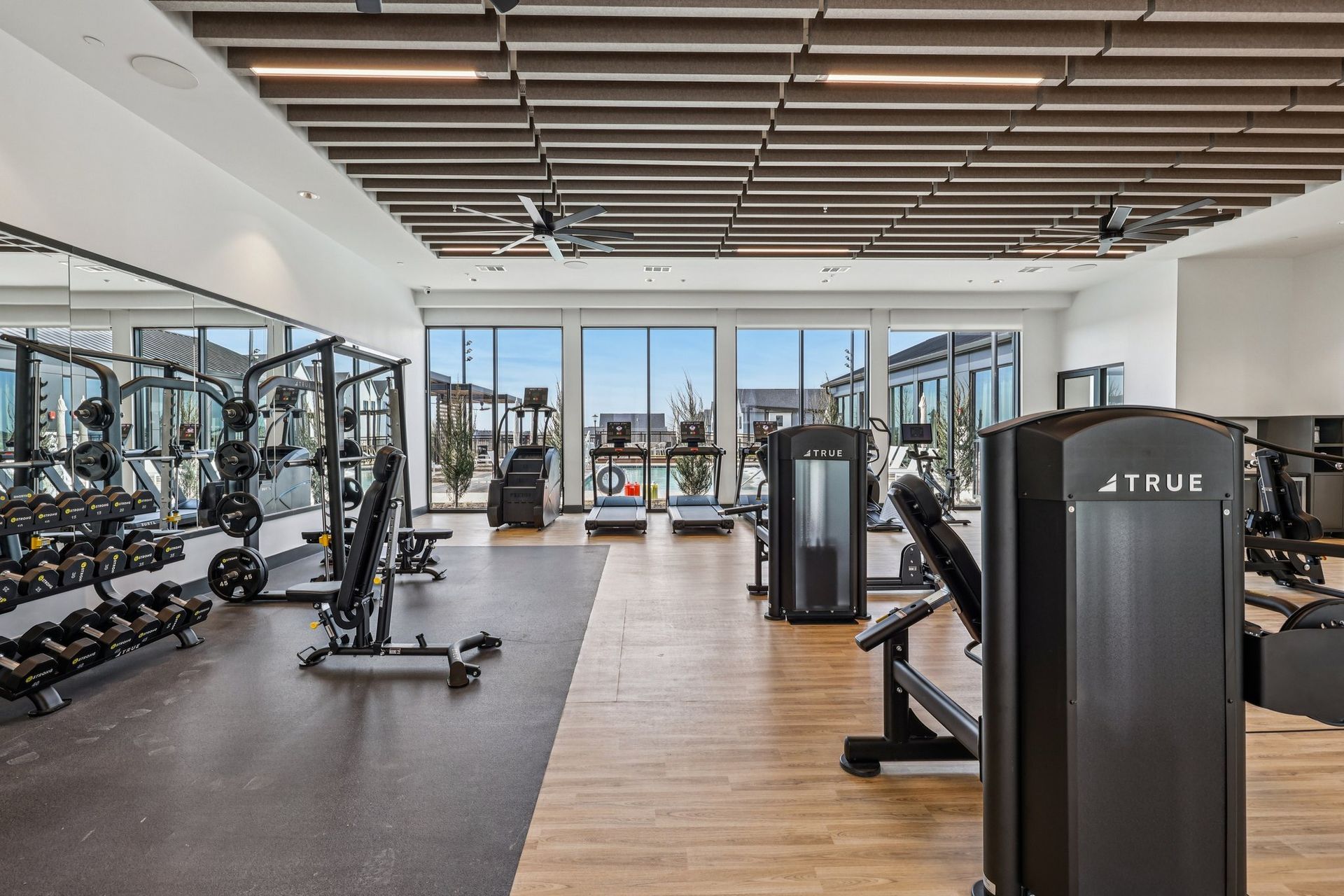 A modern, brightly lit fitness center with weight machines, free weights, and large windows overlooking a pool area.