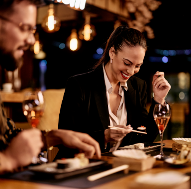 Woman enjoying sushi and a drink at a restaurant, smiling. A person is partially visible next to her.