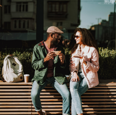 Couple eating sandwiches, smiling, seated on bench outdoors. Coffee cup and backpack nearby.