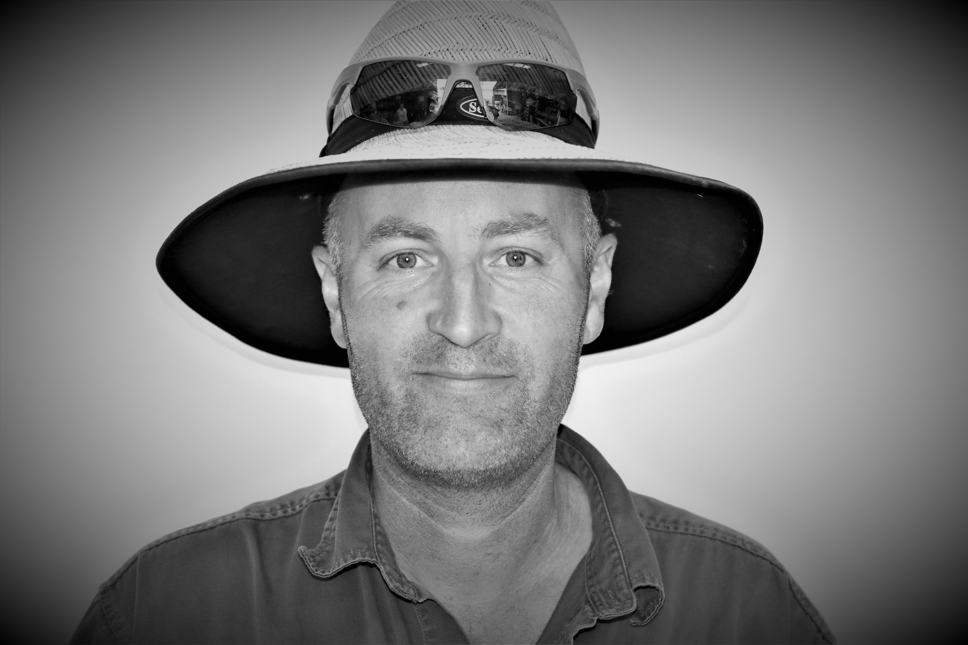 A man wearing a hat and sunglasses is smiling in a black and white photo.Image placeholder