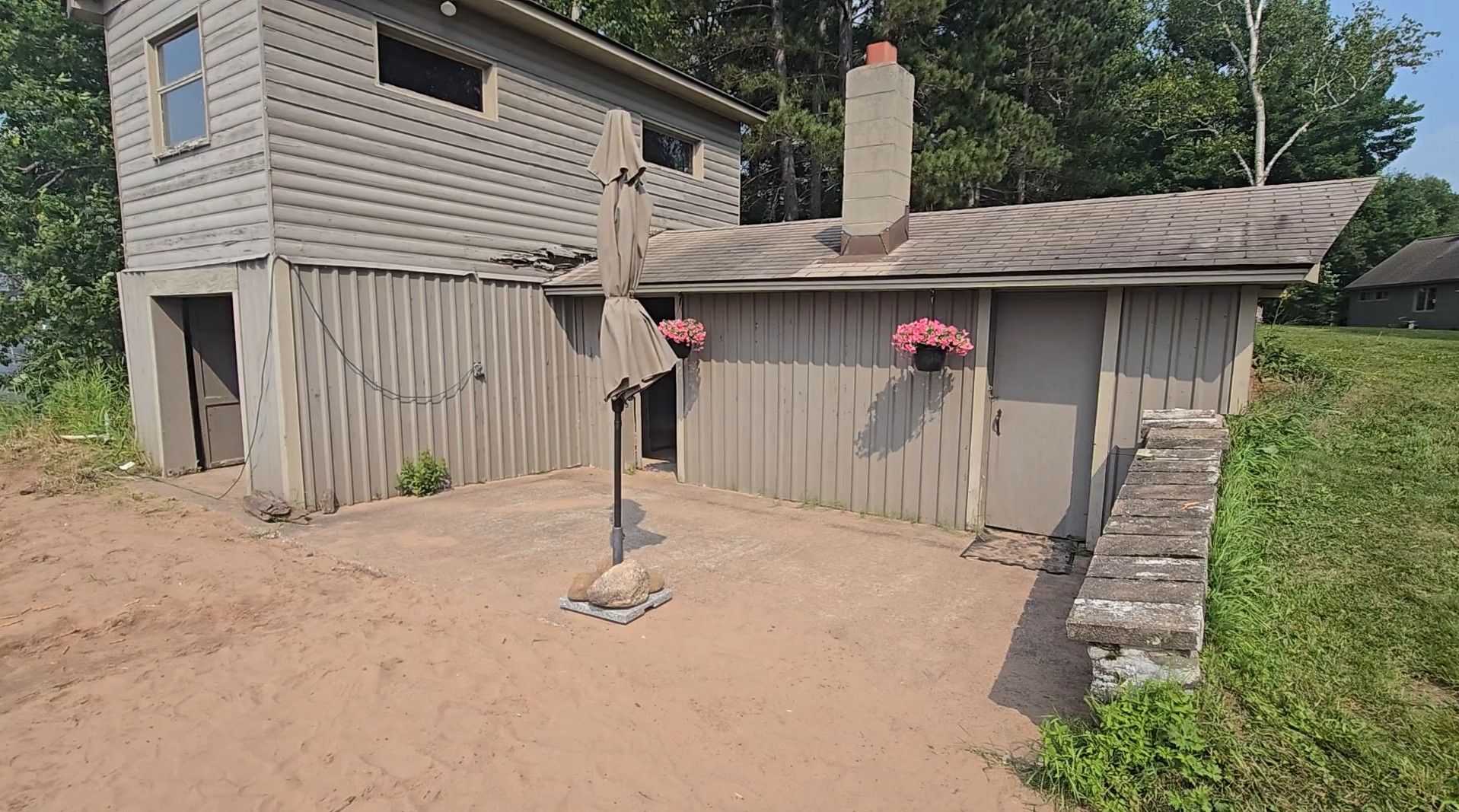 Gray wooden building with chimney, flowers, and open doorway, set on a sandy area with green grass.