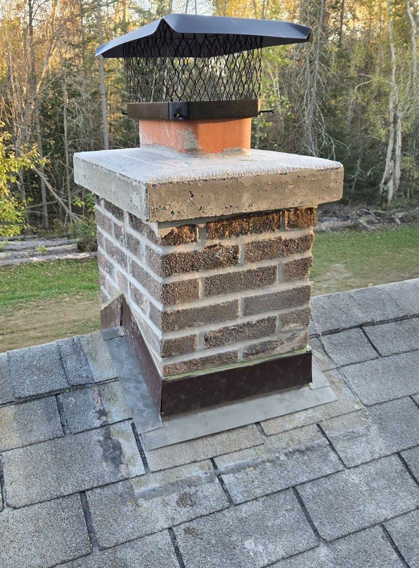 Brick chimney with concrete cap and metal chimney cap on a shingled roof.